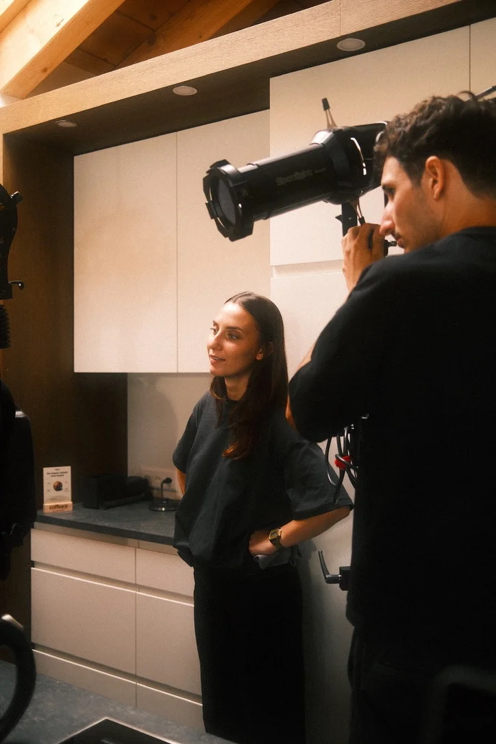 A woman standing in a kitchen being photographed by a man with a large camera.