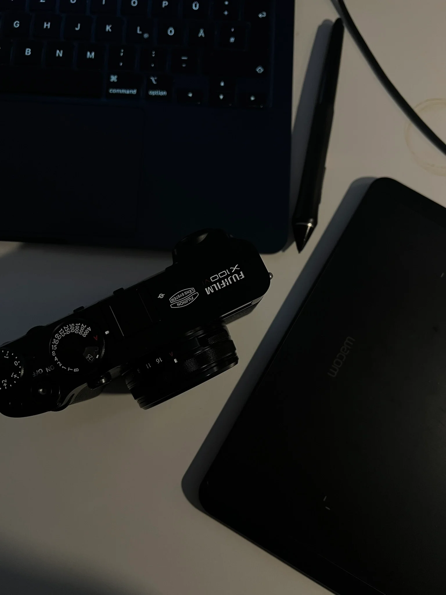 Close-up of a camera with lens markings, a black tablet, a digital pen, a laptop keyboard, and a white mouse on a white surface.
