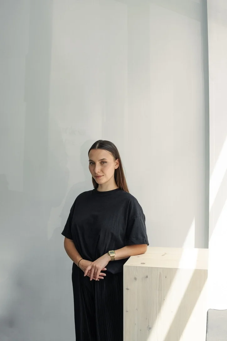 A woman with long brown hair wearing a black t-shirt and skirt, standing indoors next to a light-colored wooden surface, with natural light illuminating her from the right side.