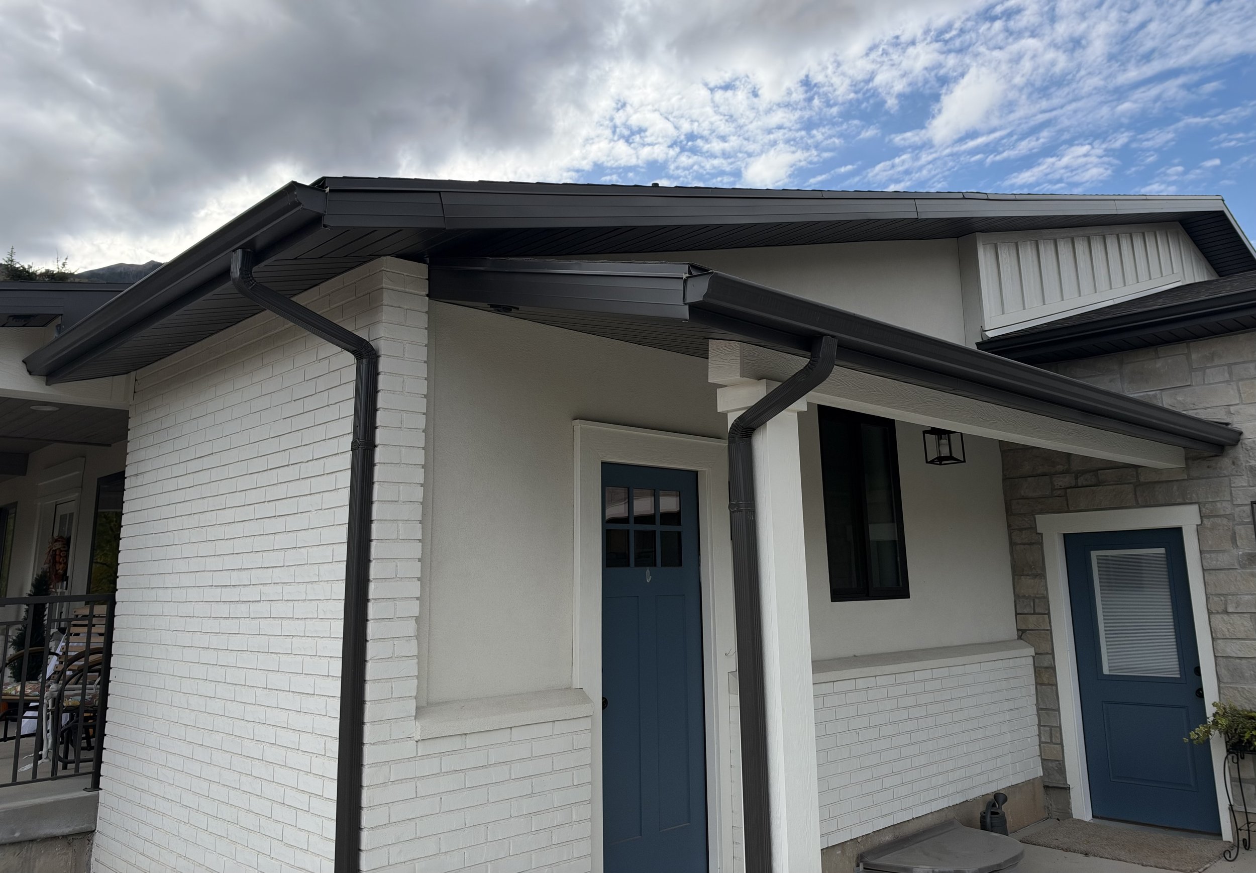 Black K-style gutters and downspouts installed on a white home with blue exterior doors and contrasting dark roof trim.