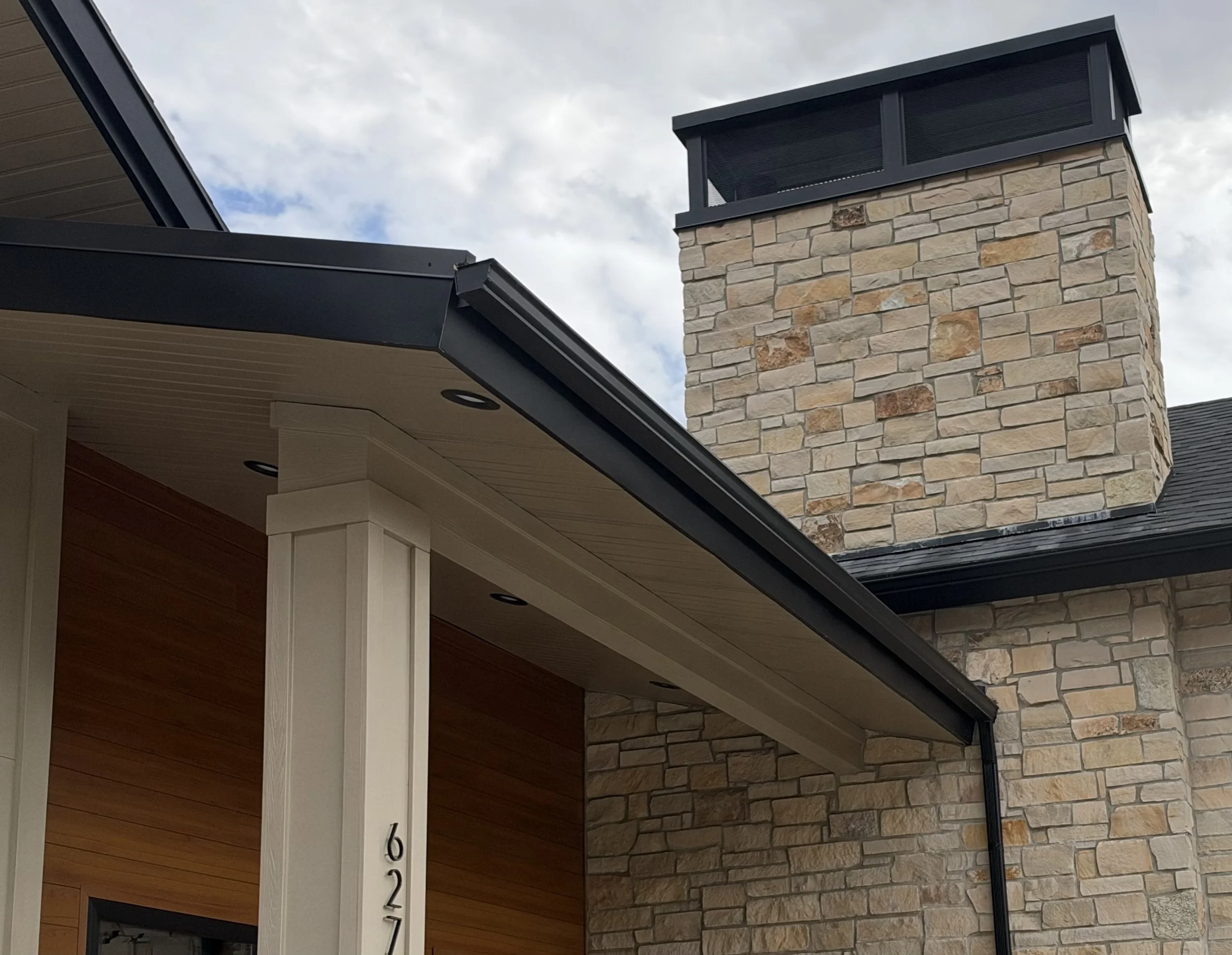 Black K-style gutter and downspout installation along a covered porch with stone masonry and dark exterior trim.