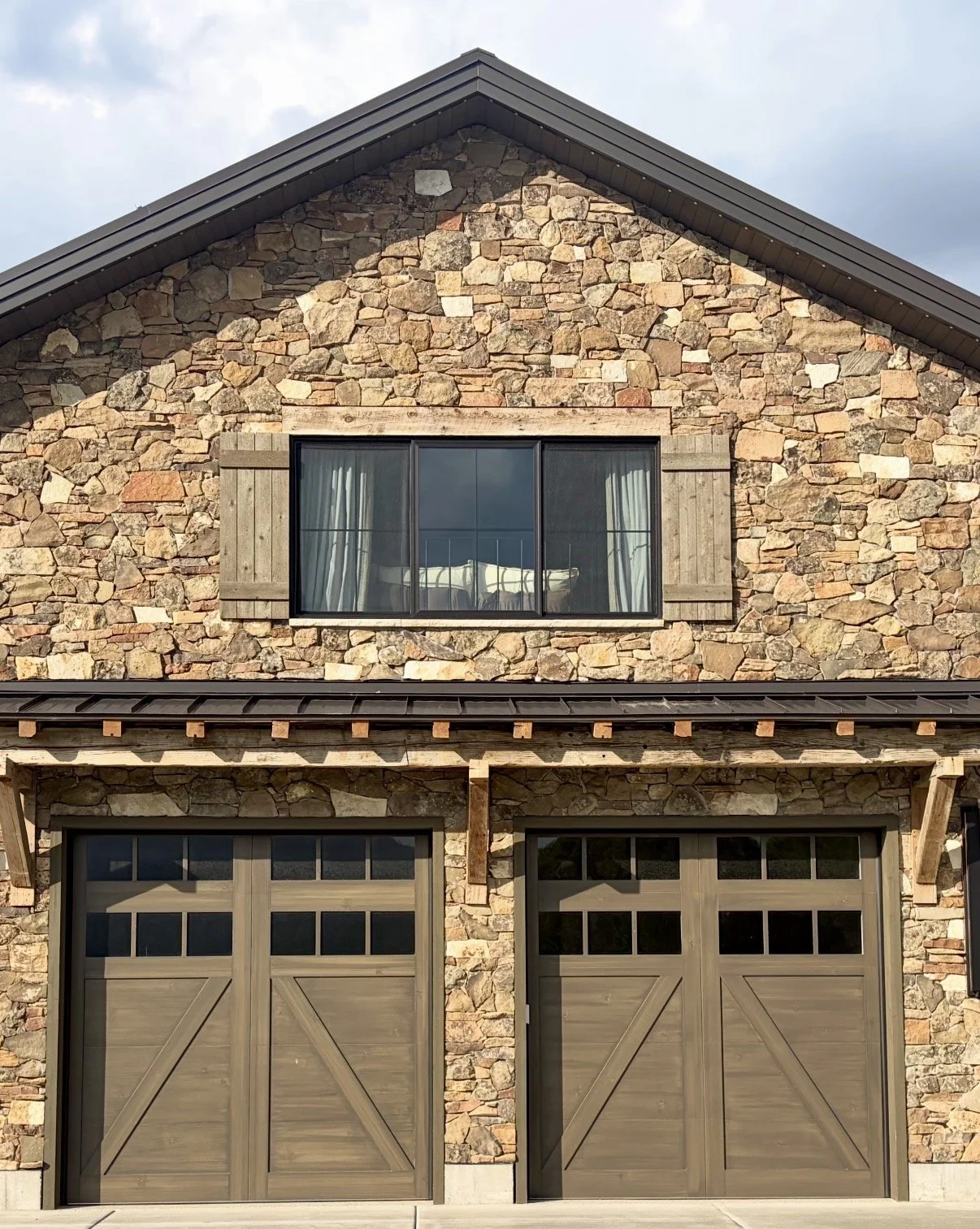 Custom wood shutters framing an upper-story window above garage doors on a stone home