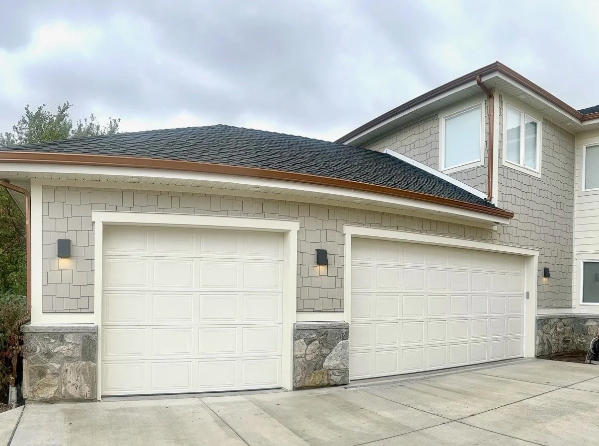 Light-colored shake-style siding on a residential garage with white trim, two garage doors, and stone accents.