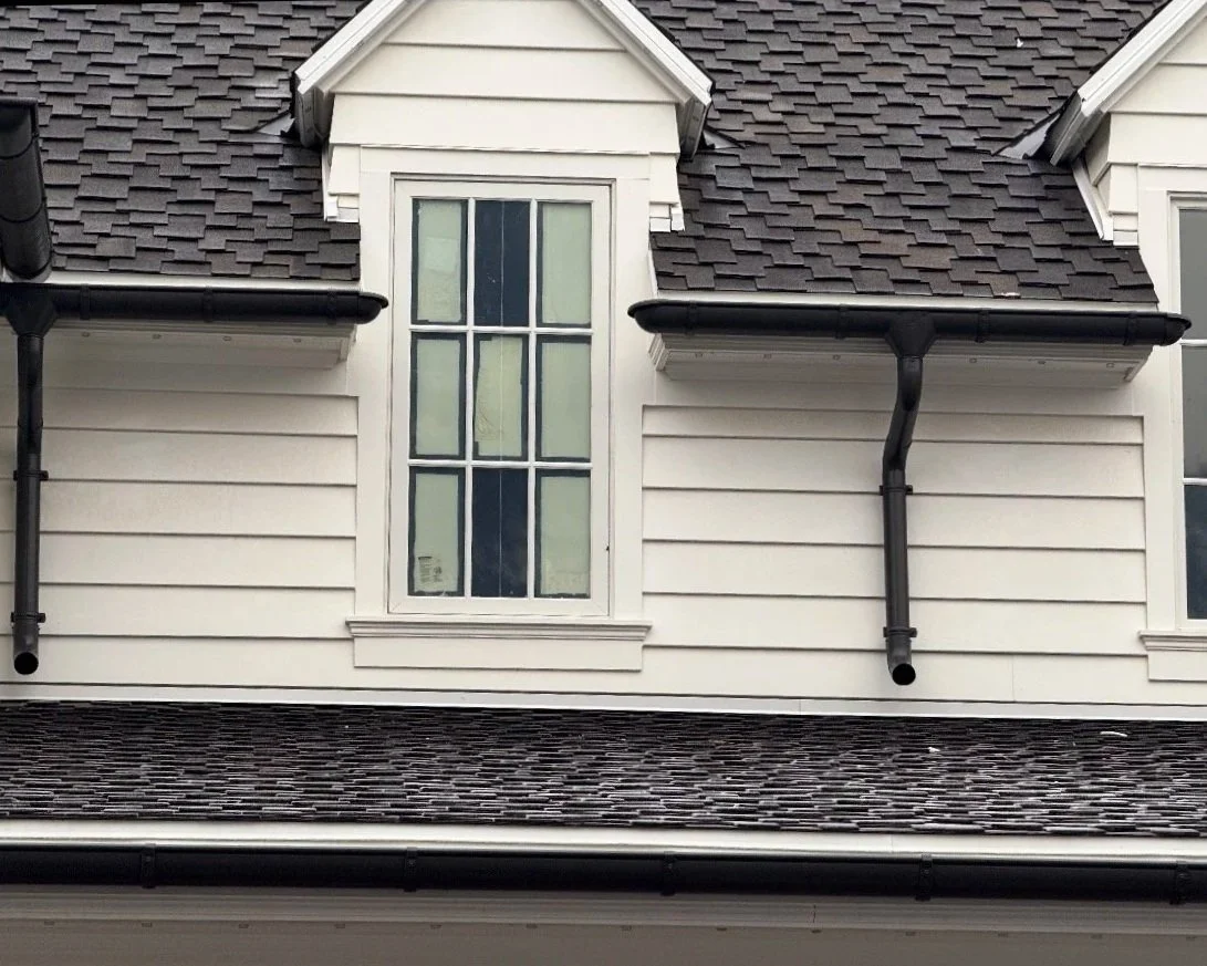 Dark half-round gutters with decorative downspouts installed on a traditional white home with dormer windows and a shingle roof.