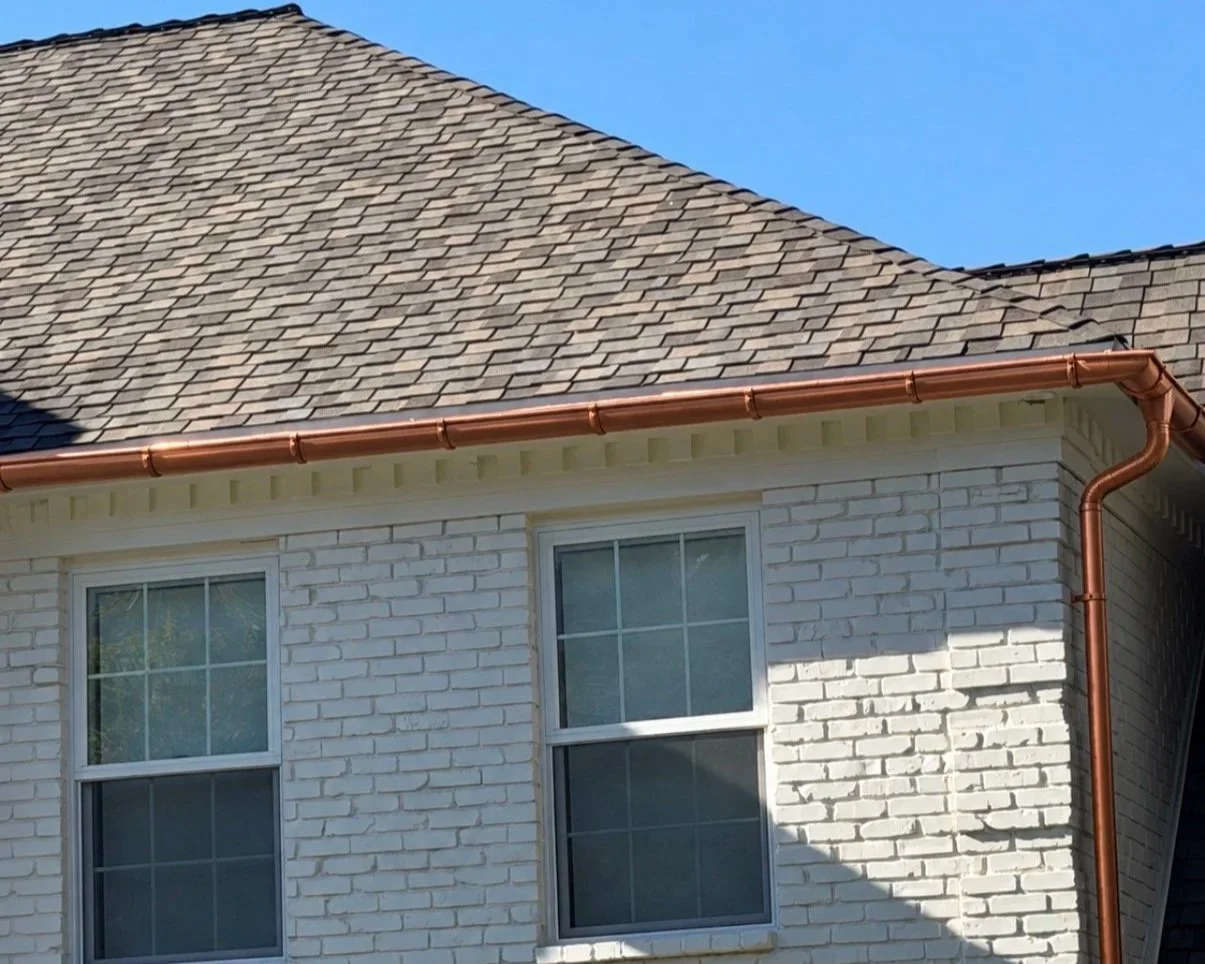 Copper half-round gutter and matching downspout installed along the roofline of a light-colored home with white brick exterior.