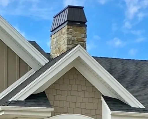 Custom dark metal chimney cap installed on a stone chimney above a residential roofline with decorative gable trim.