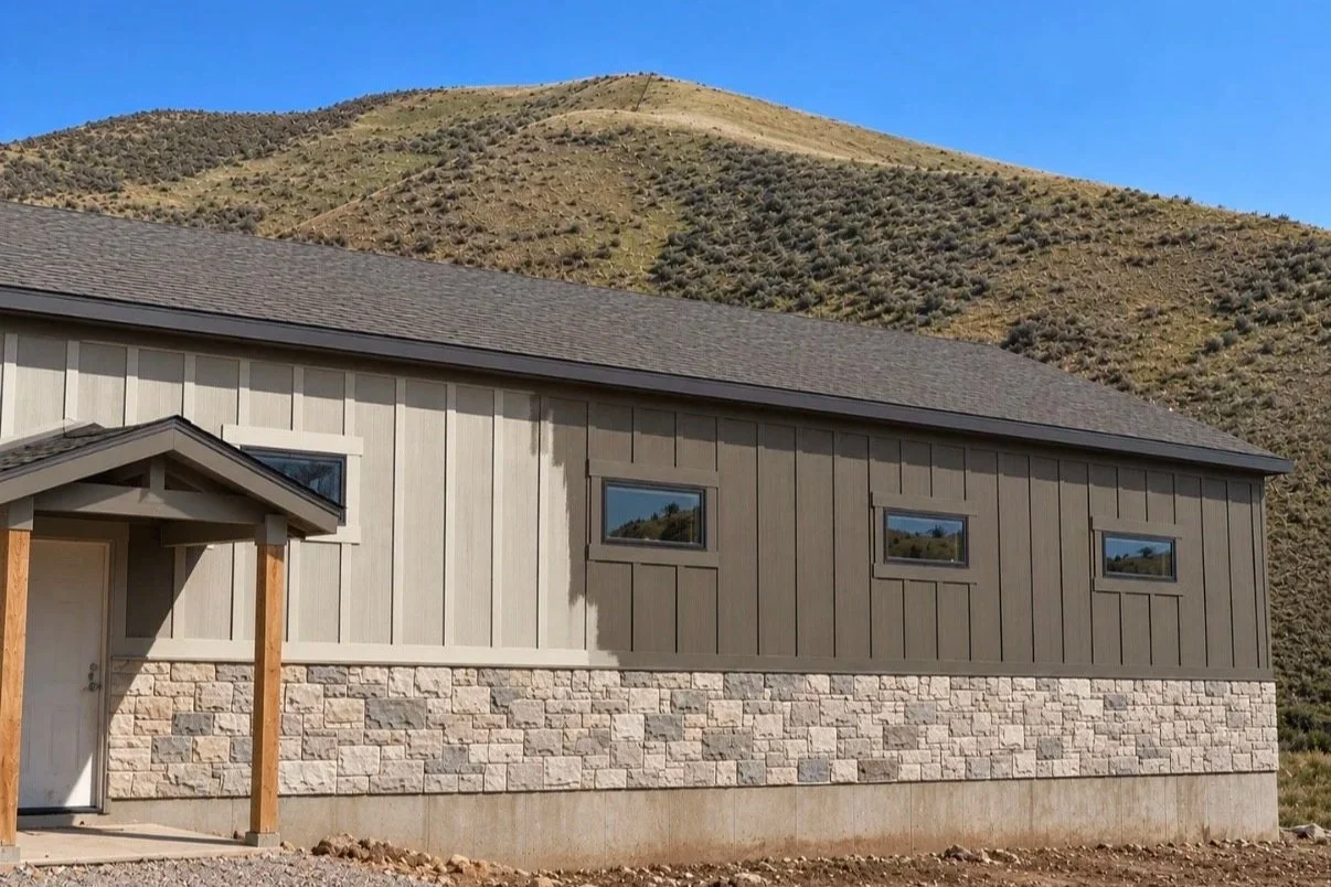 Finished exterior of a home with light-colored vertical siding, dark roof trim, and stone accents against a mountain backdrop.