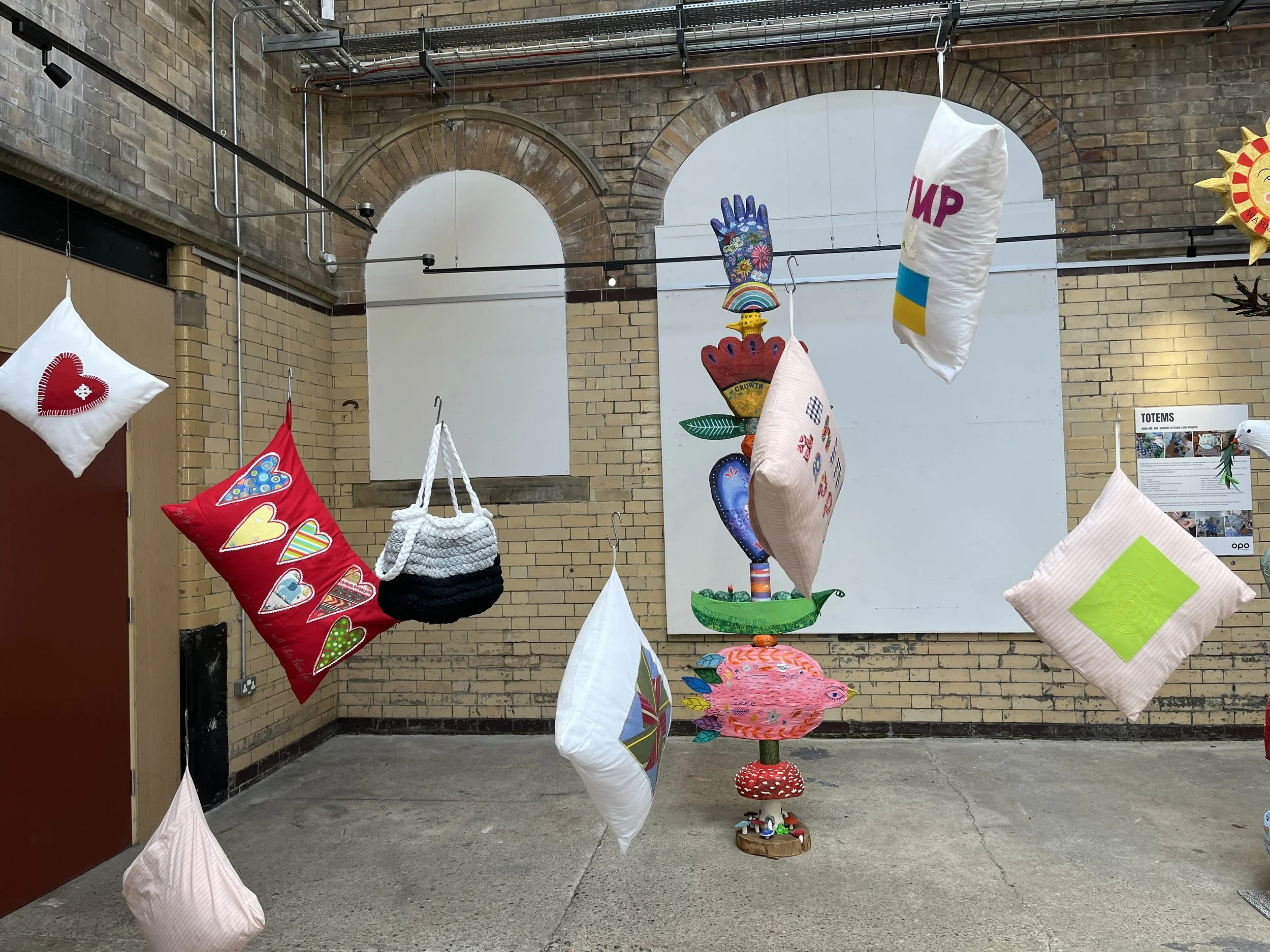Art installation featuring hanging fabric pillows and plush art pieces in a brick-walled gallery. The pillows are decorated with colorful patterns, hearts, and abstract designs, and are suspended from wires. Background includes a mural of a stylized plant with a hand-shaped top, and a notice board on the wall.