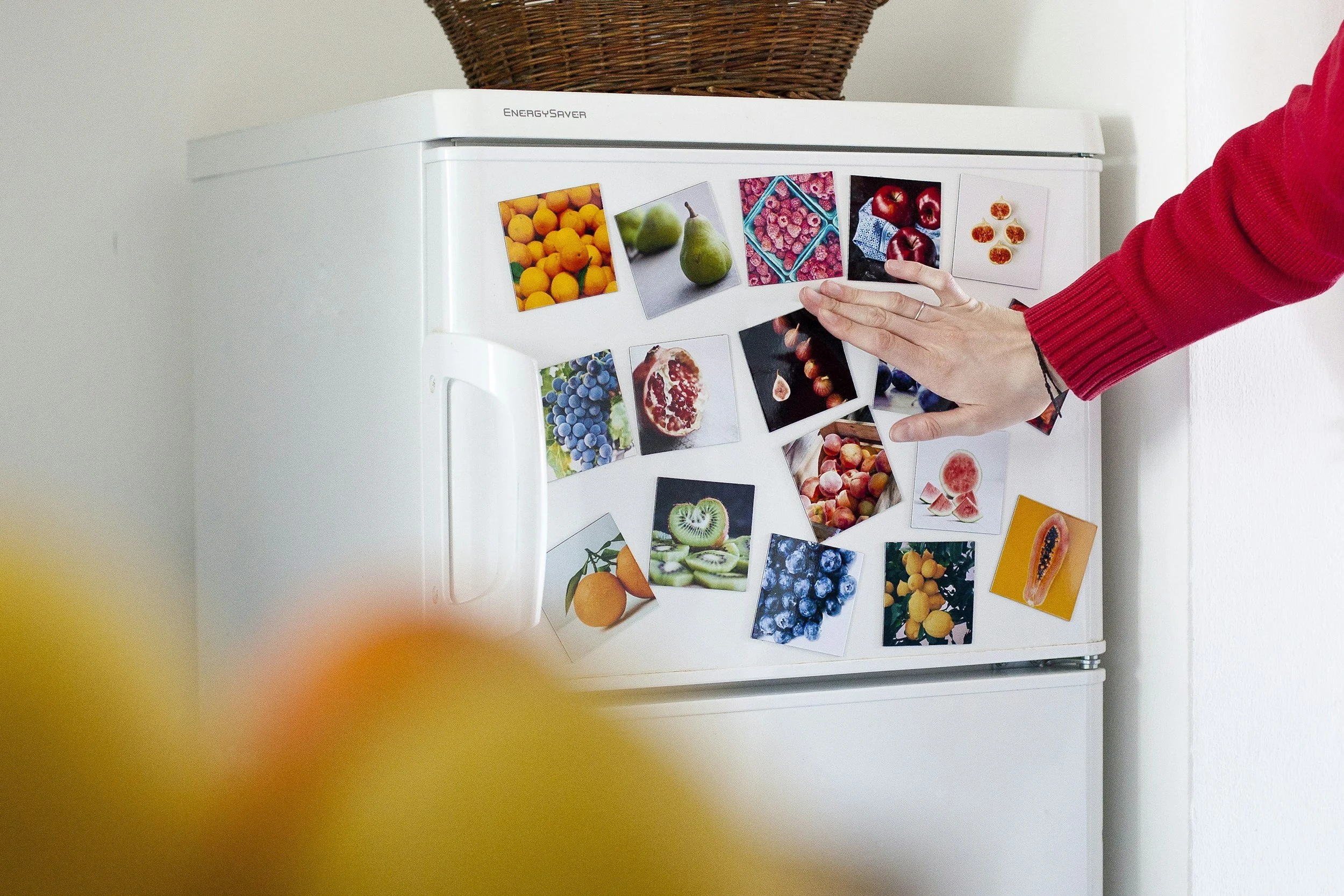 Person in red sweater placing a photo of strawberries on a white refrigerator door covered with various fruit photos.