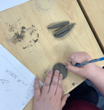 A child's hands shaping black modeling clay on a wooden table, with a pencil, paper, and more clay pieces nearby.