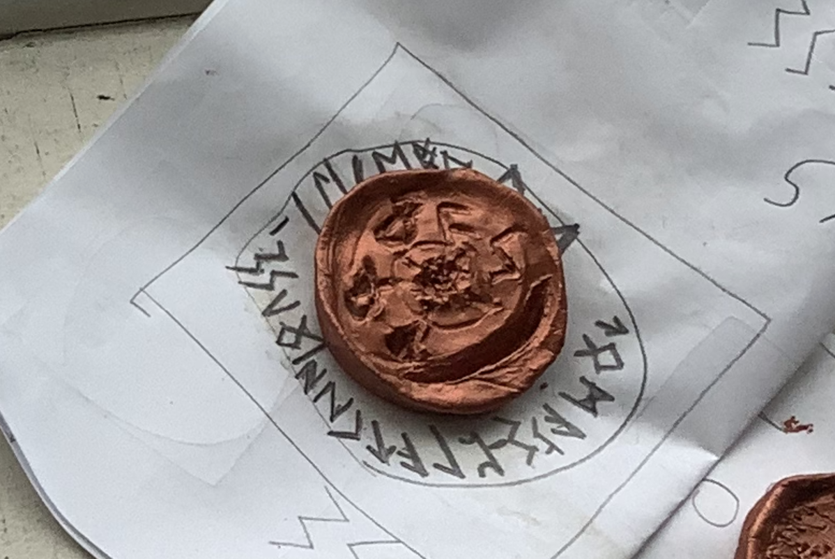 A copper-colored wax seal with a carved design placed on a piece of paper containing a circular rune or alphabet pattern.