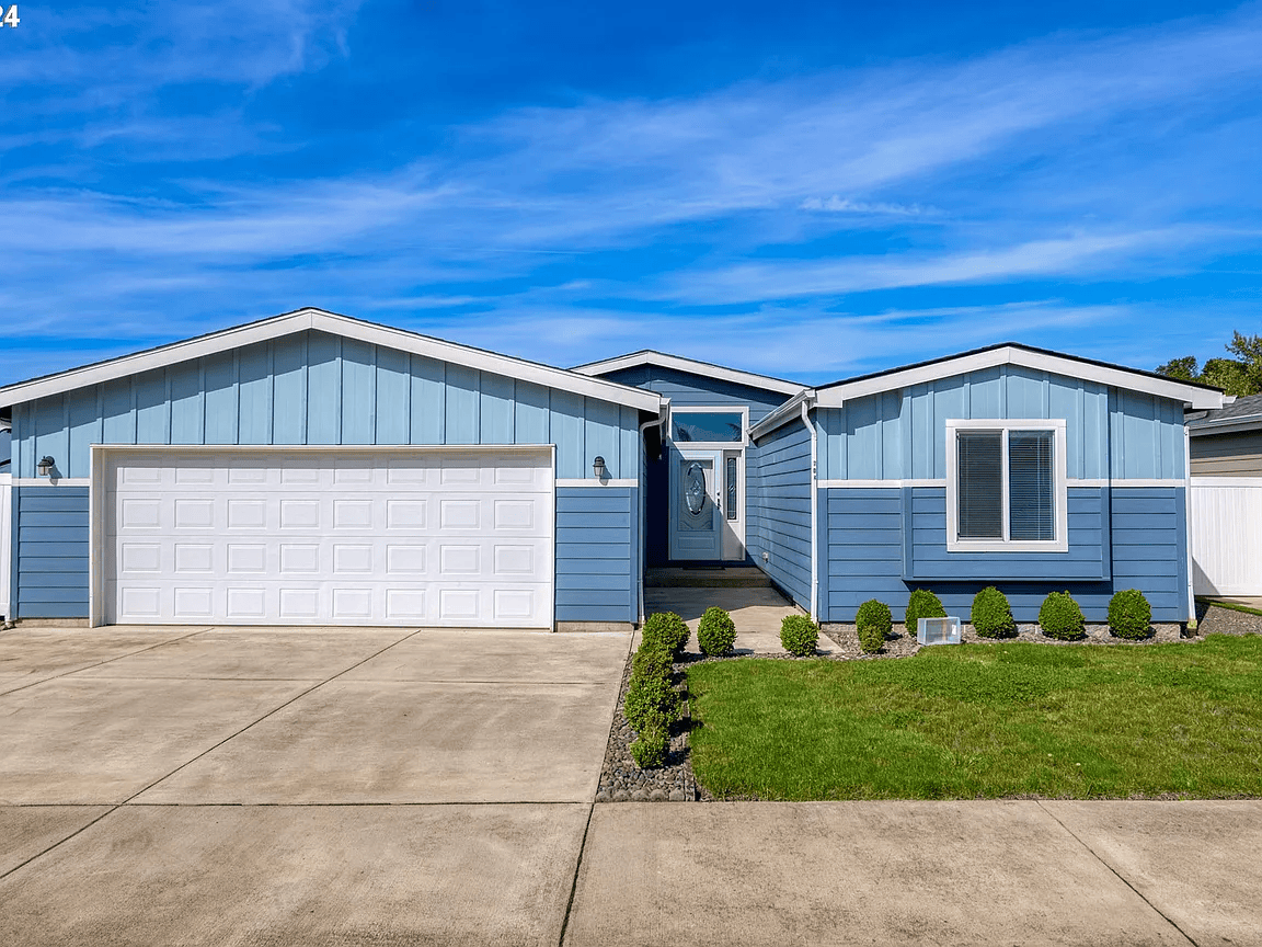 Front view of a blue house with a white garage door, shrubbery along the walkway, and a bright blue sky.