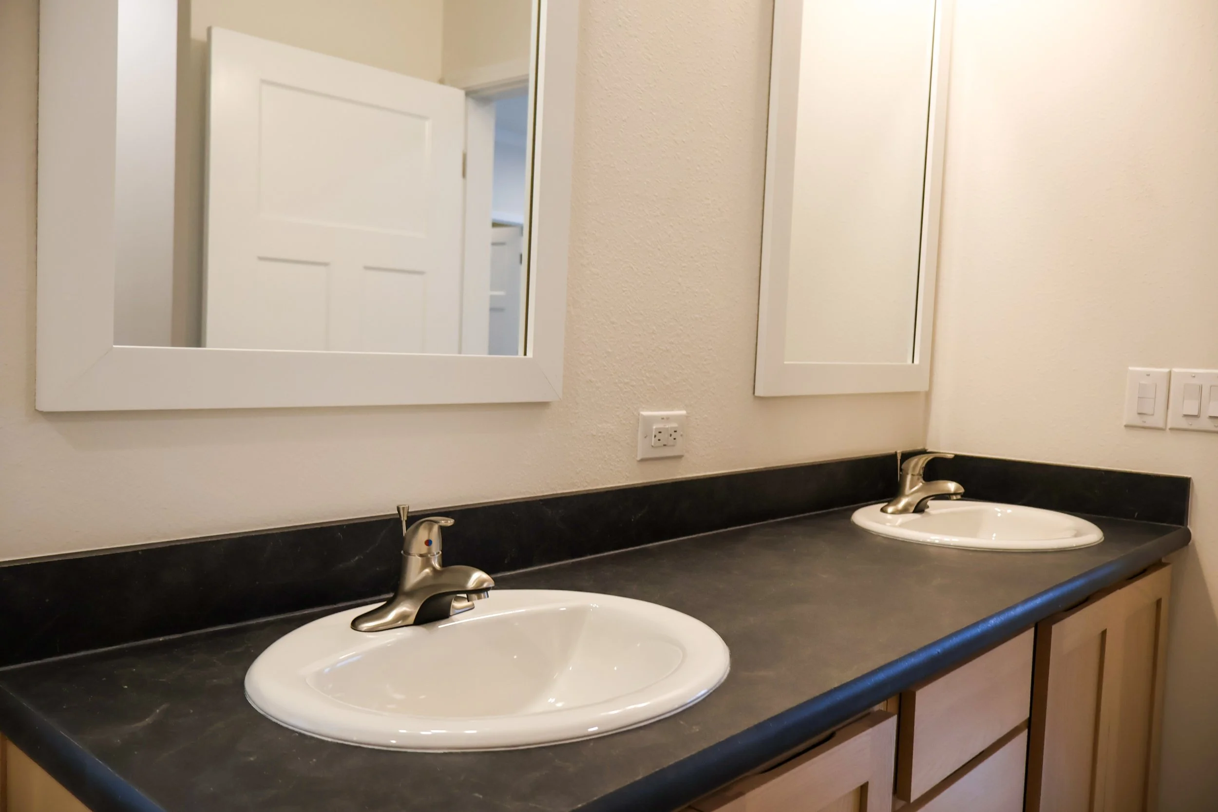 A bathroom countertop with two sinks and two mirrors above. The countertop is black, with wooden cabinet beneath. The background shows a partially open door and neutral-colored walls.
