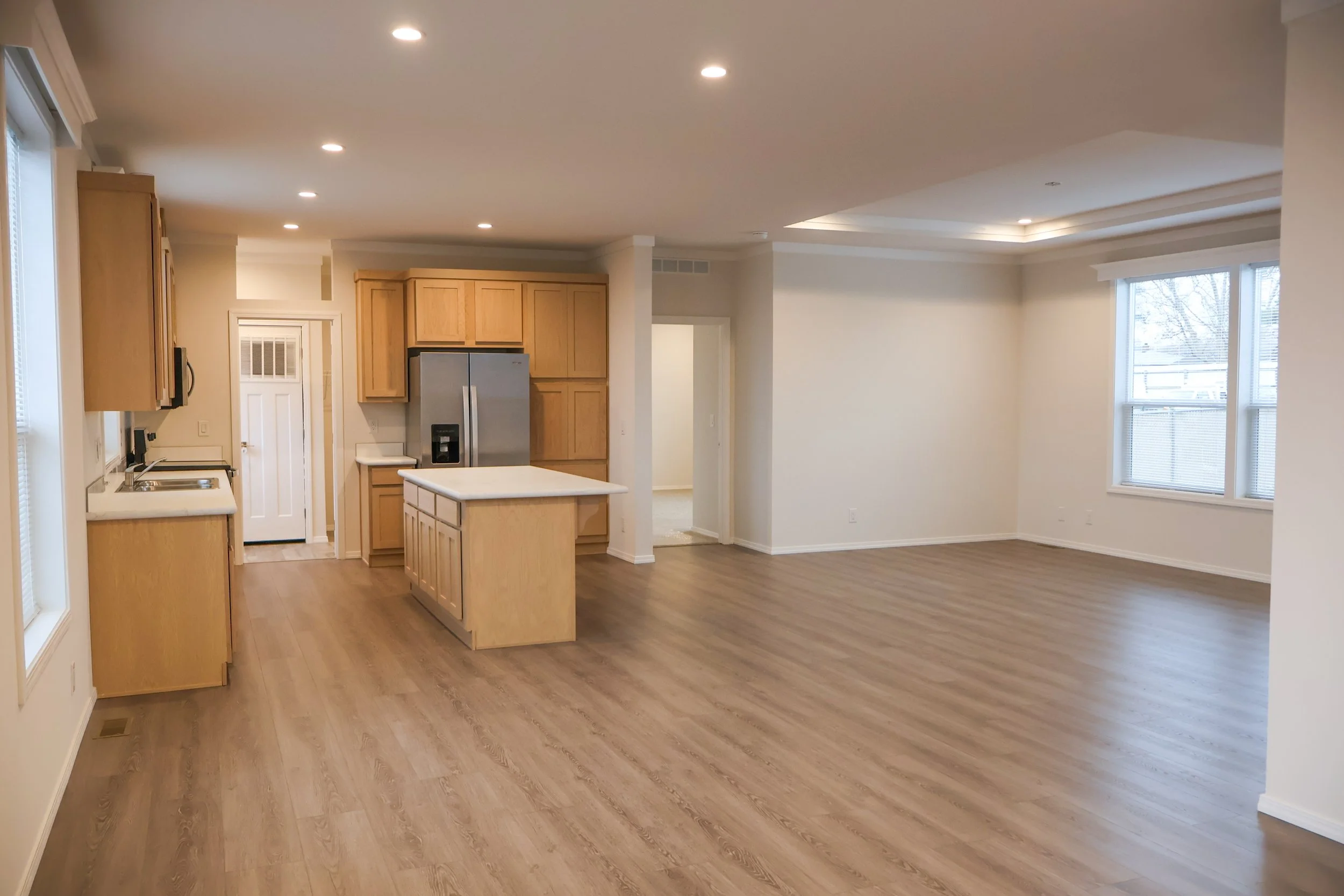 Empty open-concept living room and kitchen with light wood flooring, wooden cabinets, stainless steel refrigerator, and large windows.