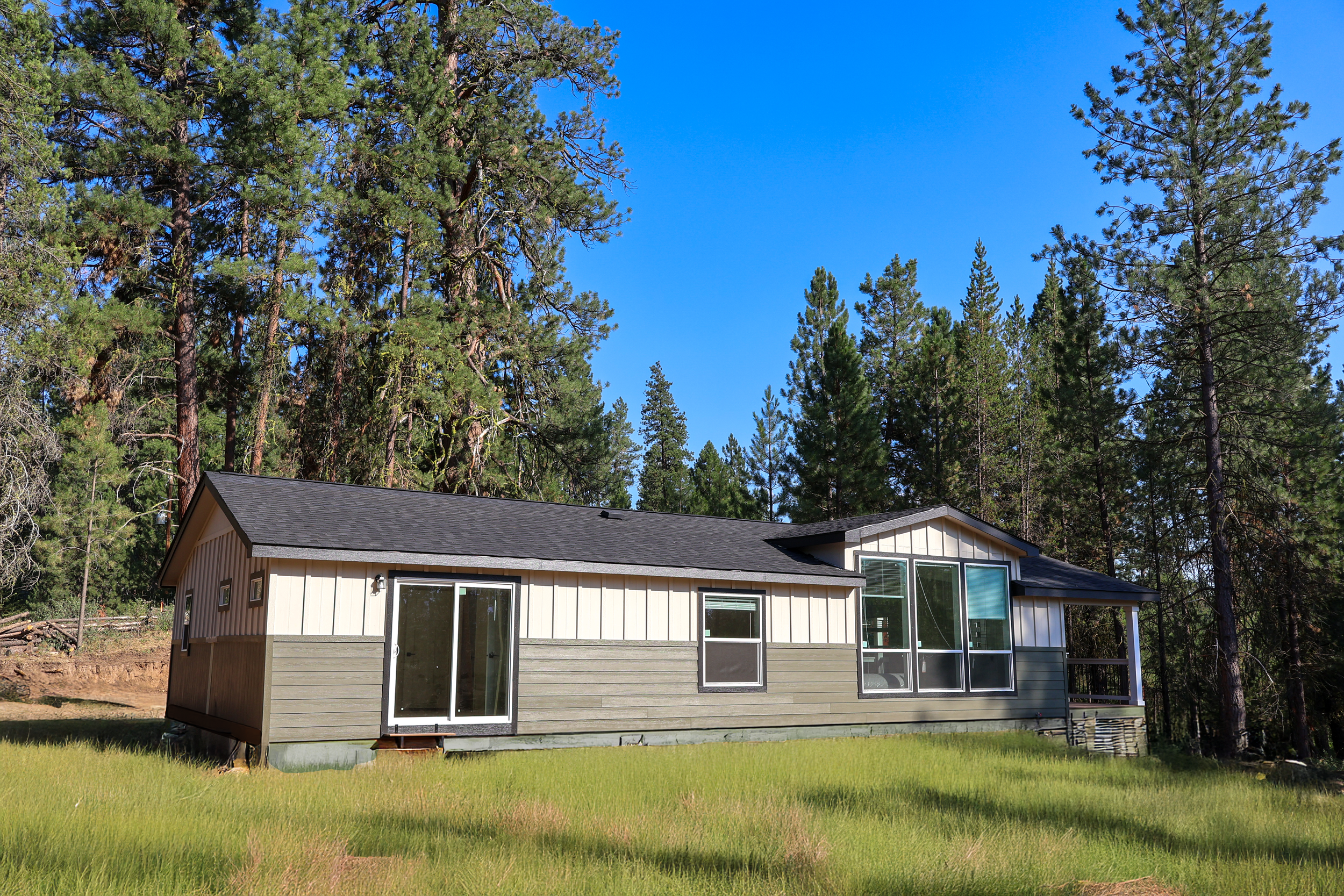 A house with glass sliding doors and large windows is surrounded by green grass and tall pine trees under a clear blue sky.