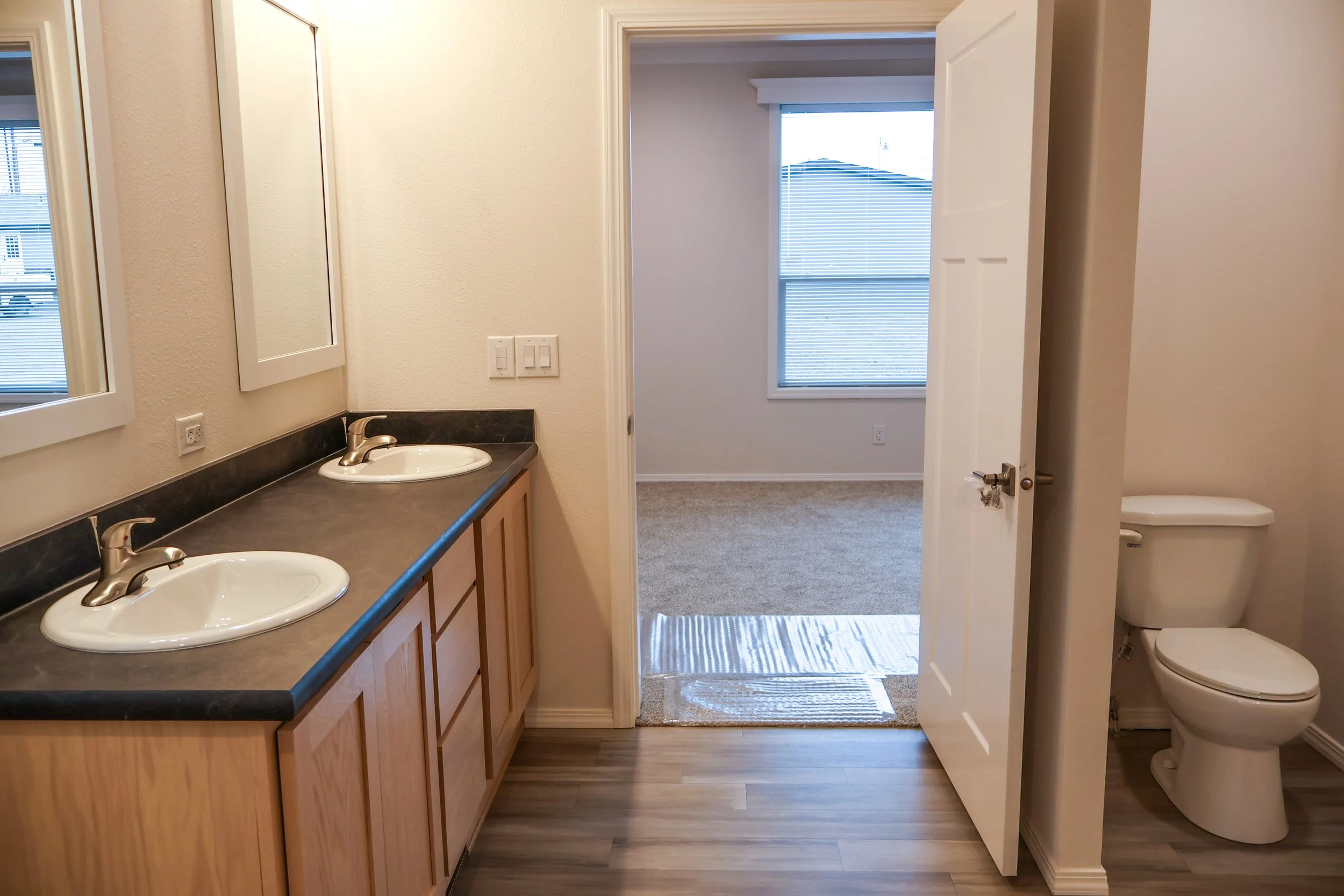 Bathroom with dual sinks, mirrors, and a separate toilet area, adjacent to a carpeted room with window blinds.