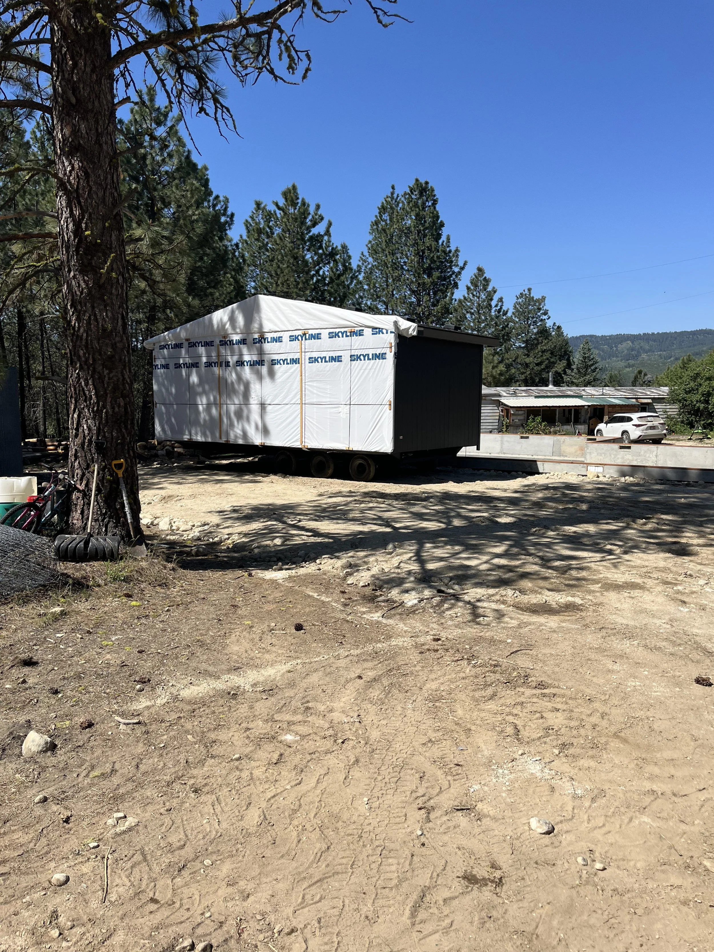 A construction site with a trailer wrapped in white weather-resistant material labeled 'Skyline' parked on a dirt area surrounded by tall pine trees and a partially constructed building in the background.
