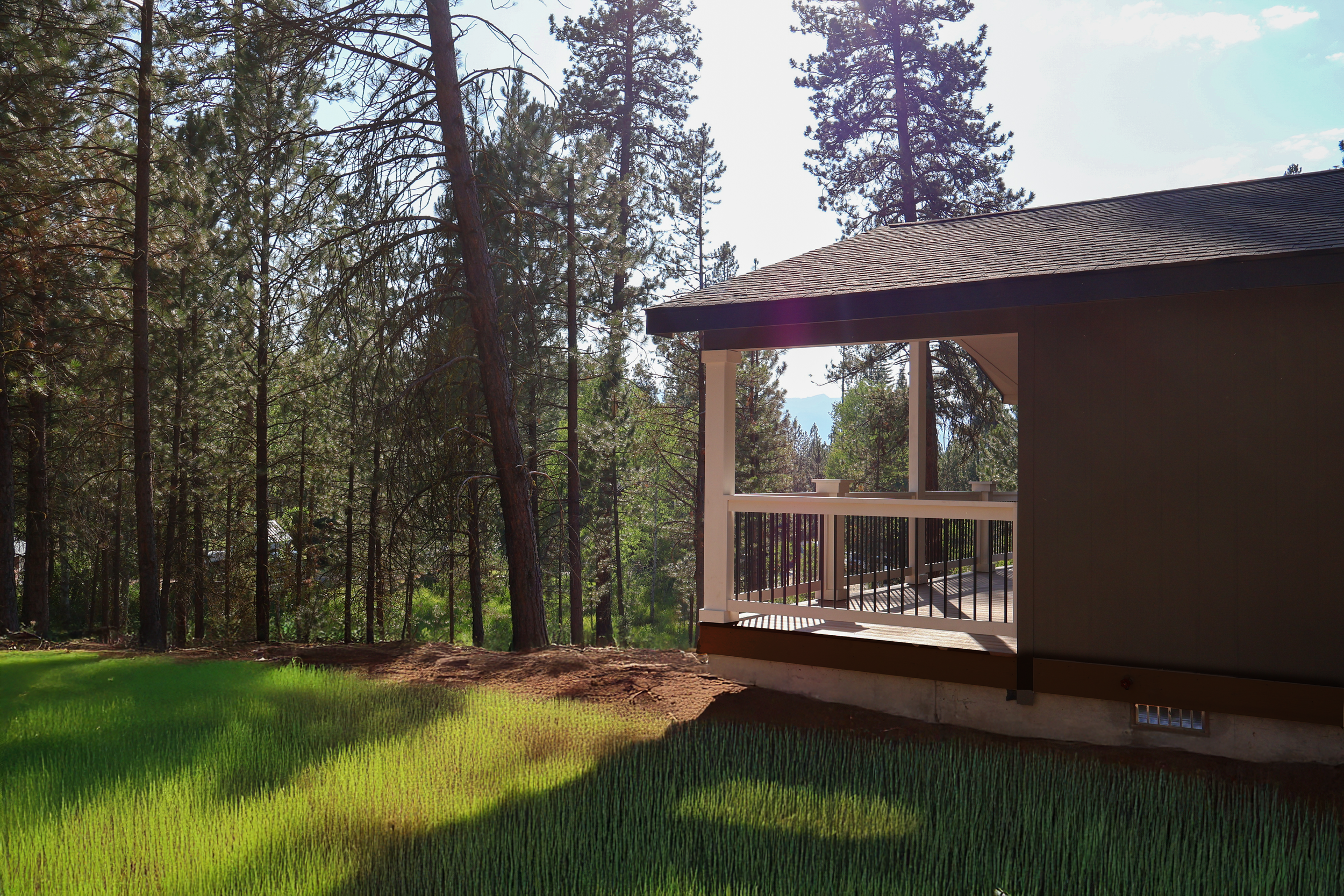 A house with a porch overlooking a forest with tall pine trees, and a green lawn in the foreground under a partly cloudy sky.