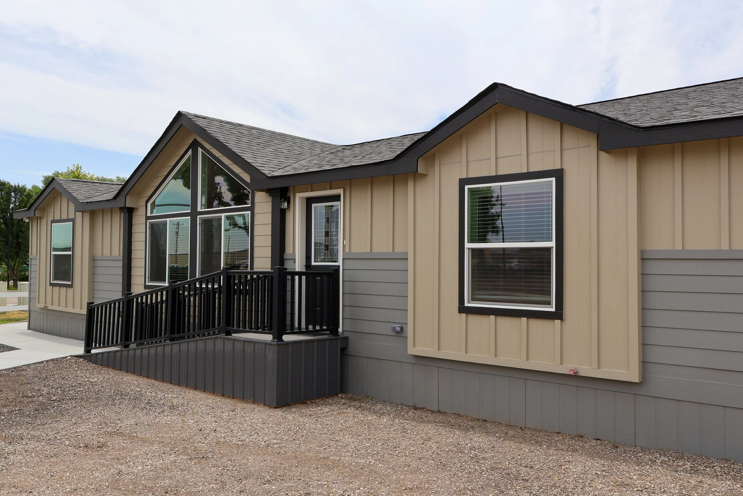 Exterior of a modern house with beige vertical siding, gray lower siding, black window frames, and a small black porch with railings.
