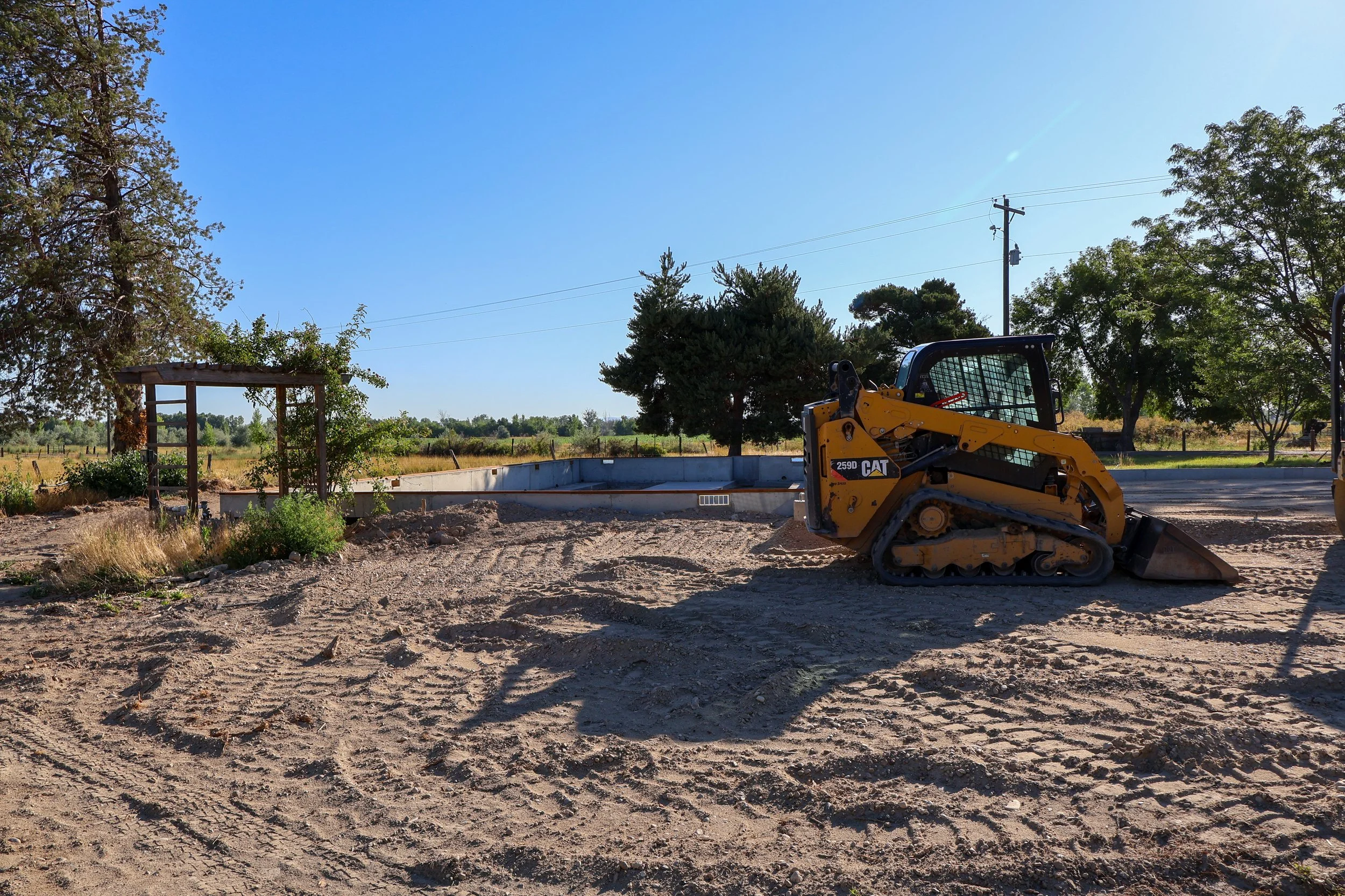 A small yellow Caterpillar compact loader on a dirt construction site with trees and a clear blue sky in the background.