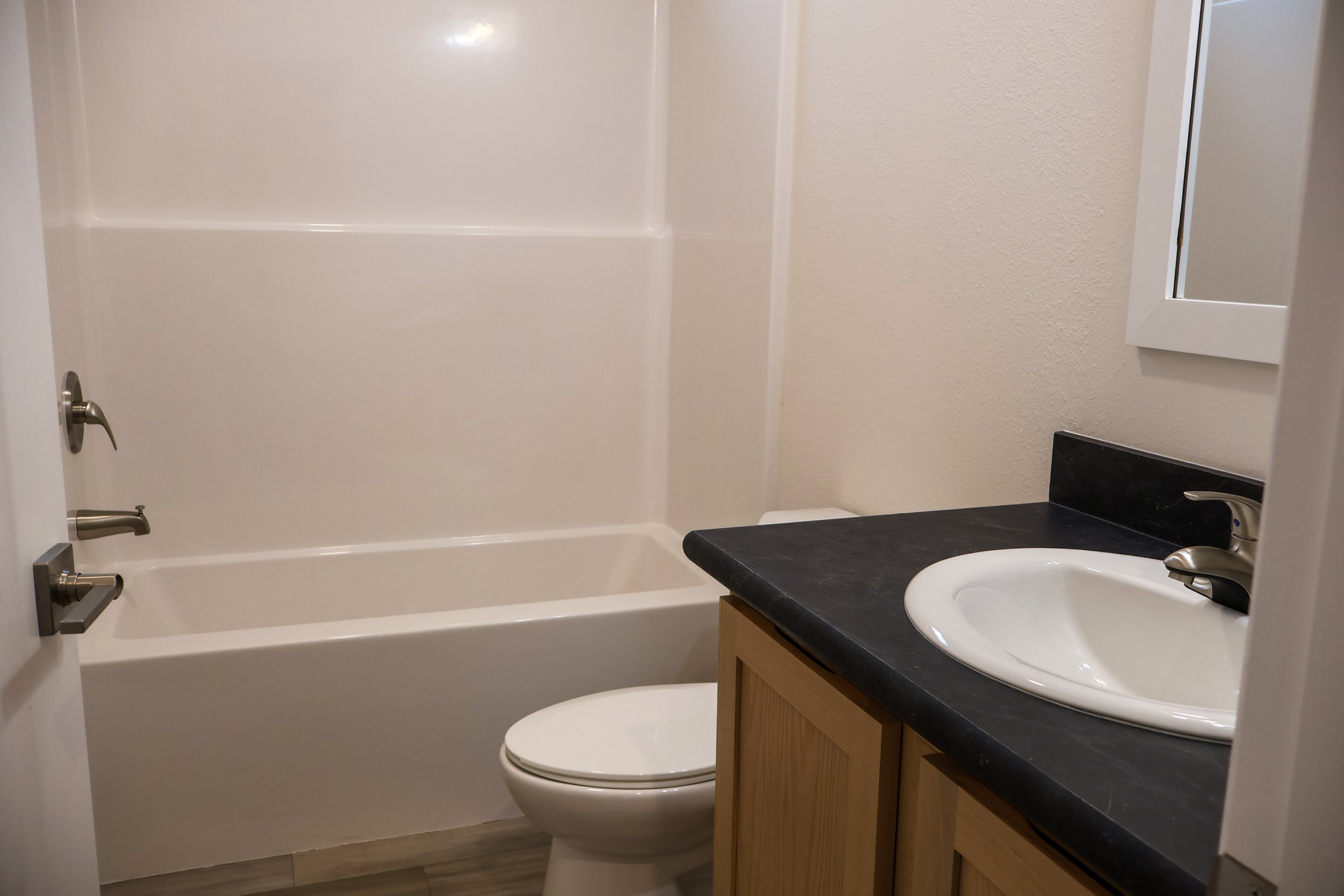 Bathroom with a bathtub, toilet, and a sink with a black countertop and a wooden cabinet, mirror on the wall.