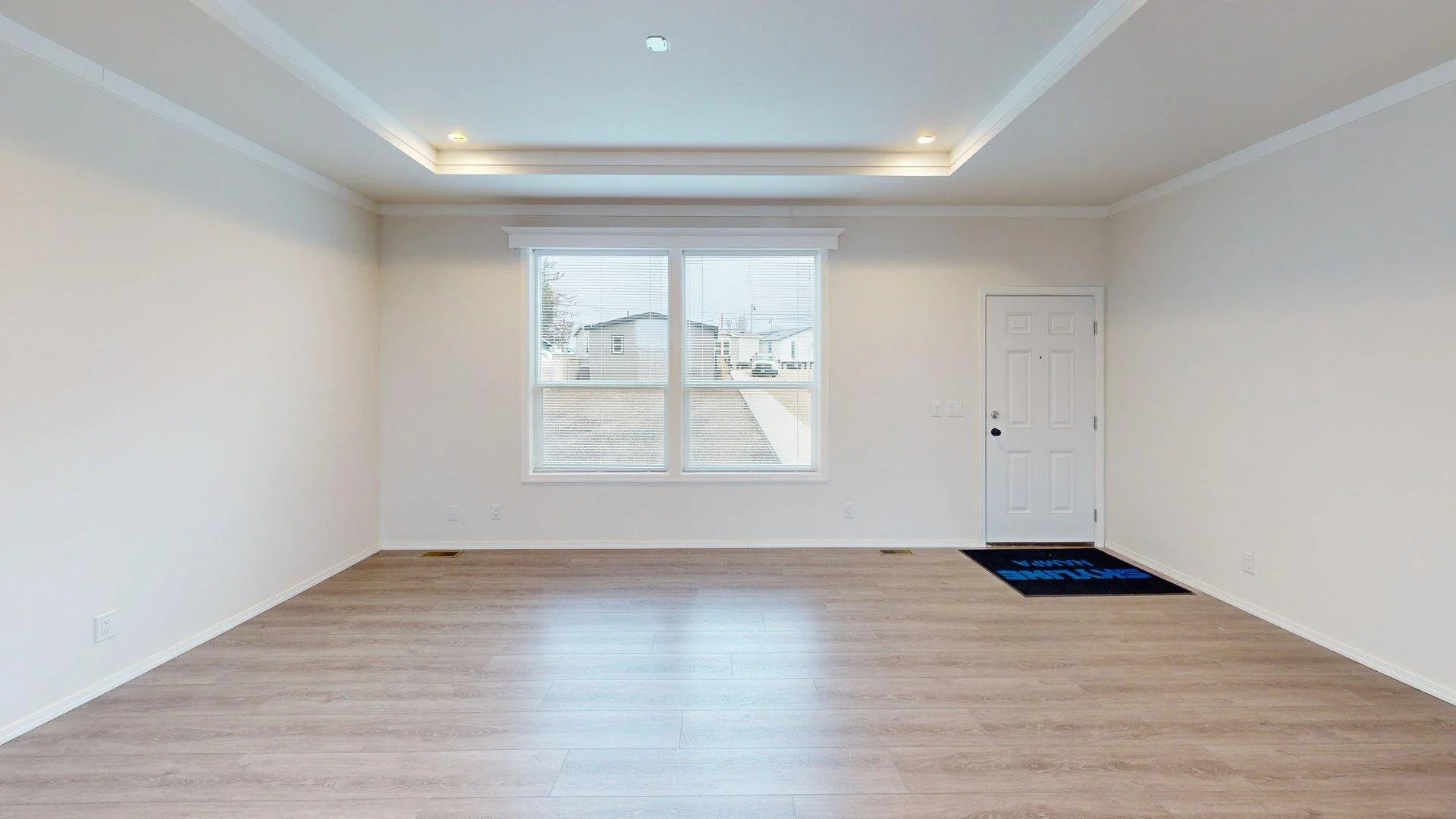 Empty living room with white walls, large window, hardwood flooring, and a front door with a welcome mat.