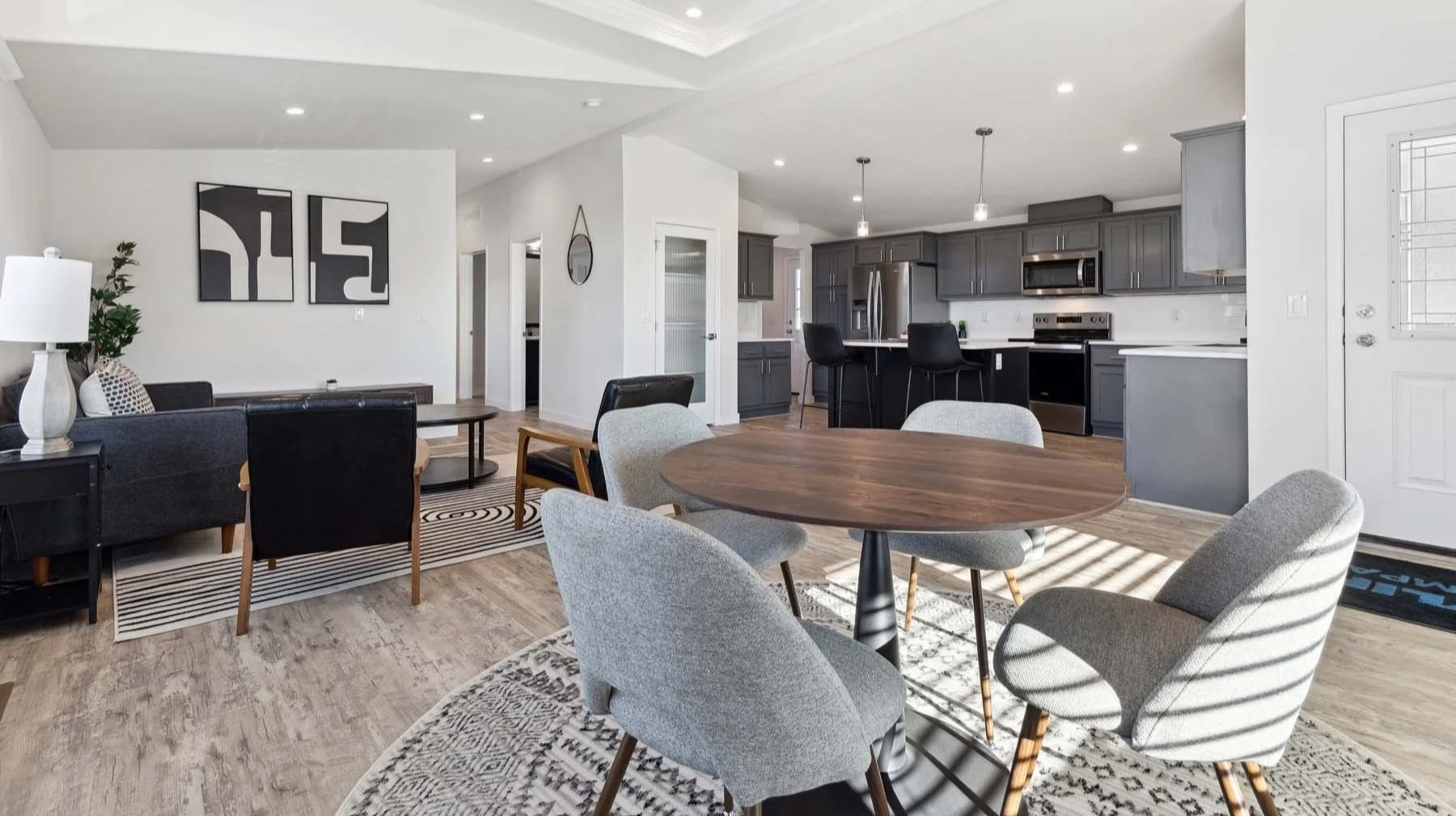 Open-concept living and dining area with a kitchen in the background. The dining table has six gray upholstered chairs, and the living area features a black sofa, a wooden coffee table, and modern wall art. The kitchen has gray cabinets, stainless steel appliances, and a breakfast bar with black stools.