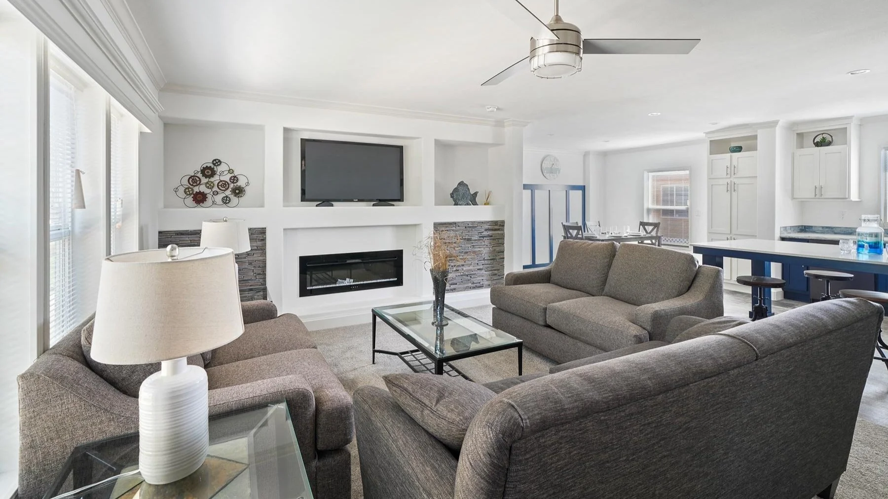 Living room with gray sofas, glass coffee table, wall-mounted TV, modern fireplace, and open kitchen with white cabinets and blue accents.