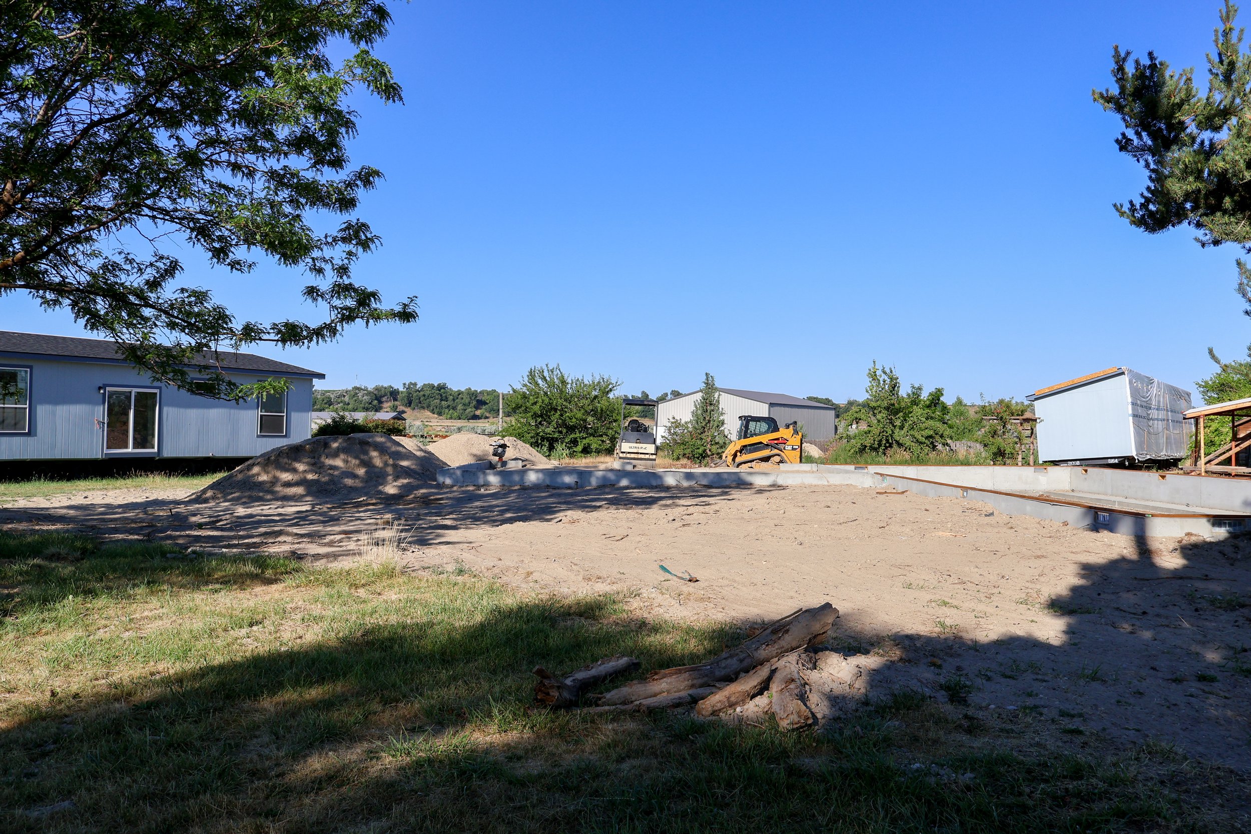 A construction site with a partially built foundation, construction equipment, a dirt pile, and surrounding trees under a clear blue sky.