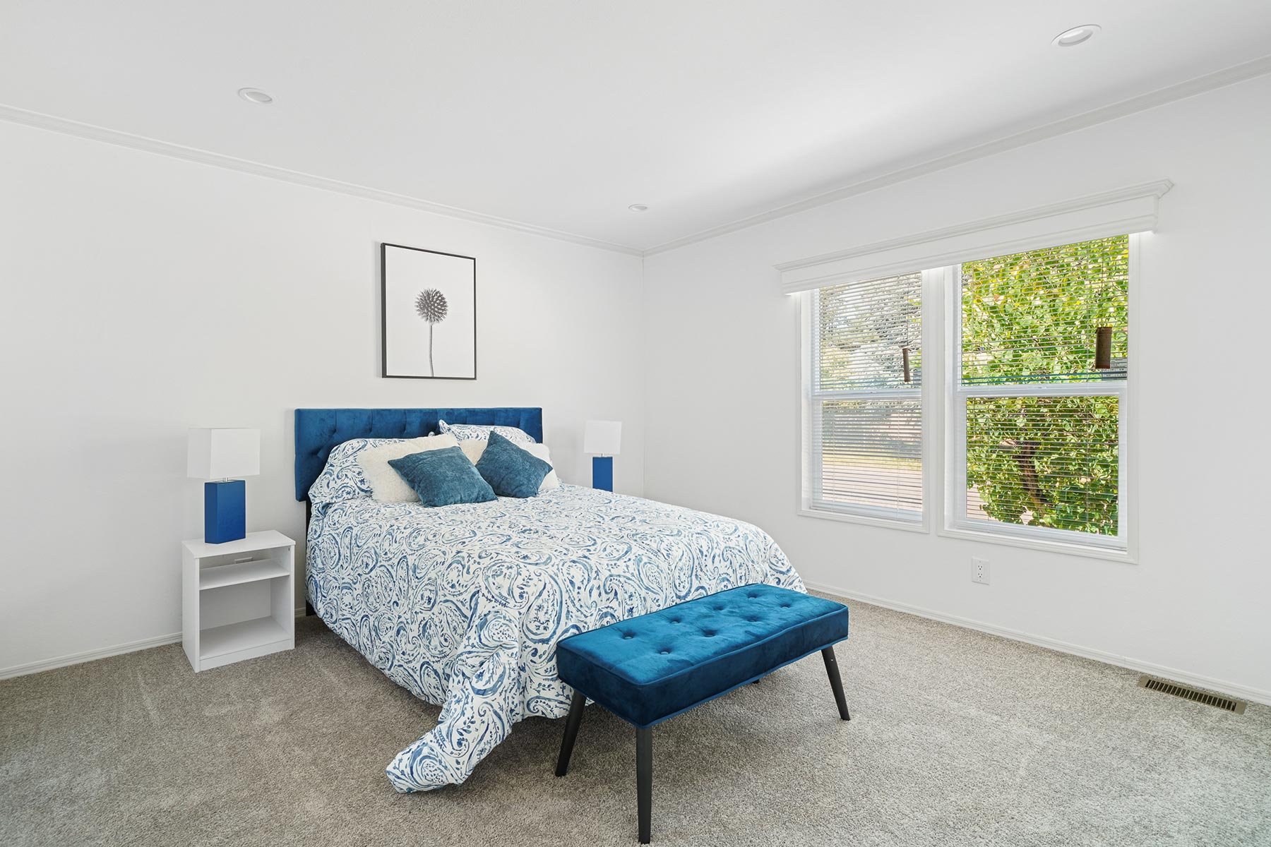 A bedroom with a white wall, a bed with a blue upholstered headboard, a patterned blue and white bedspread, and blue pillows. There are two white nightstands with white lamps, a blue upholstered bench at the foot of the bed, and a window with blinds