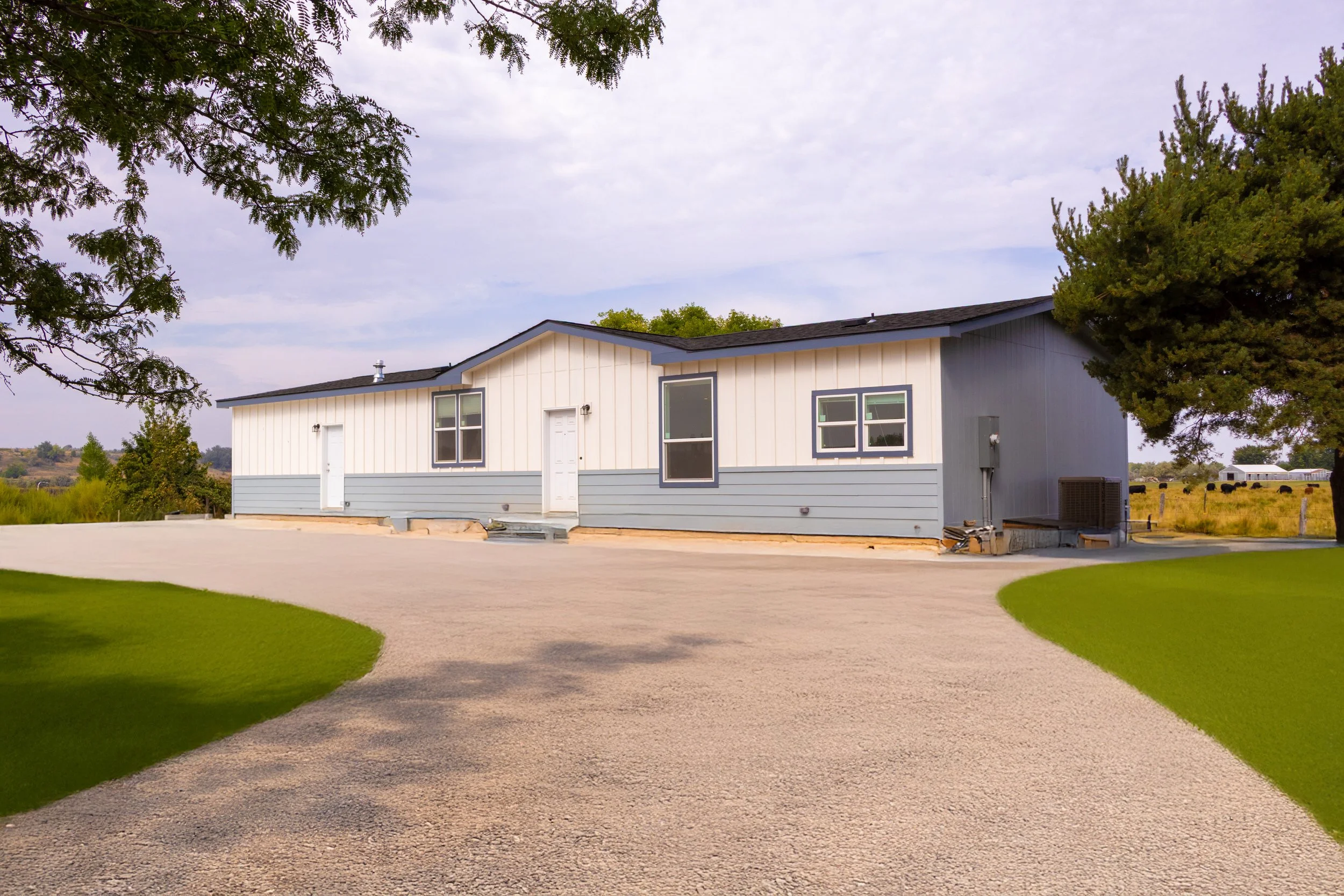A newly constructed single-story house with white and gray exterior siding, three windows and a white door, situated on a gravel driveway with green grass on either side, under a partly cloudy sky.