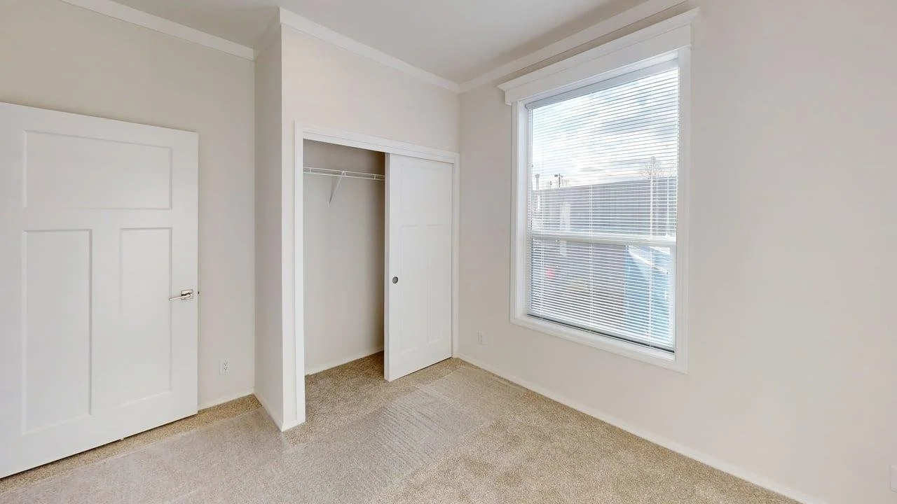 Empty room with closet and window, beige carpet and white walls in a house.