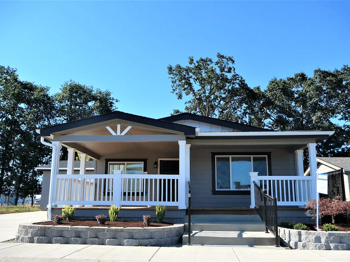 Front view of a house with a porch, white railing, and steps, surrounded by trees on a sunny day.