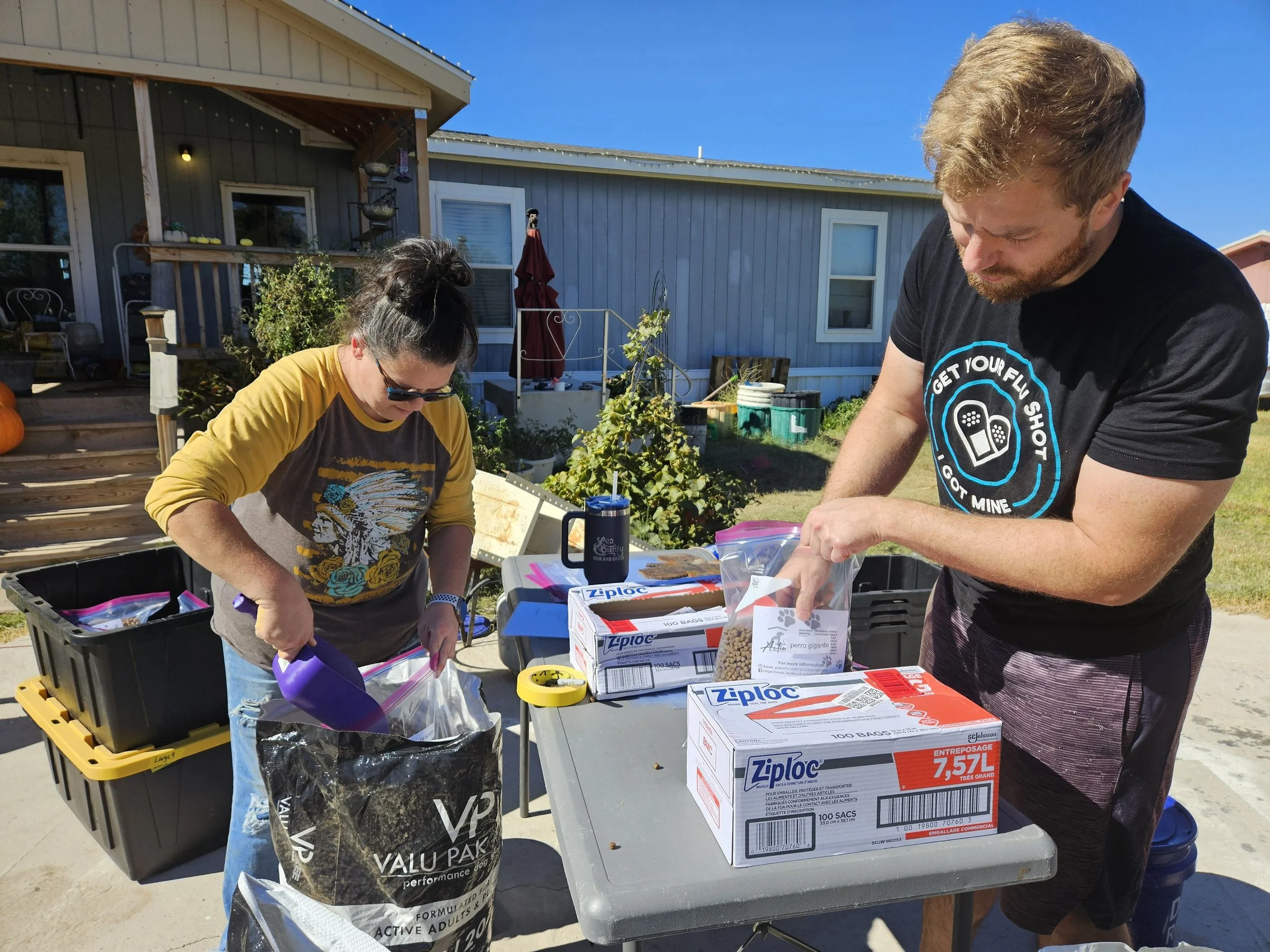 Two people outdoors, working with large storage bags, Ziploc boxes, and pet food supplies on a table.