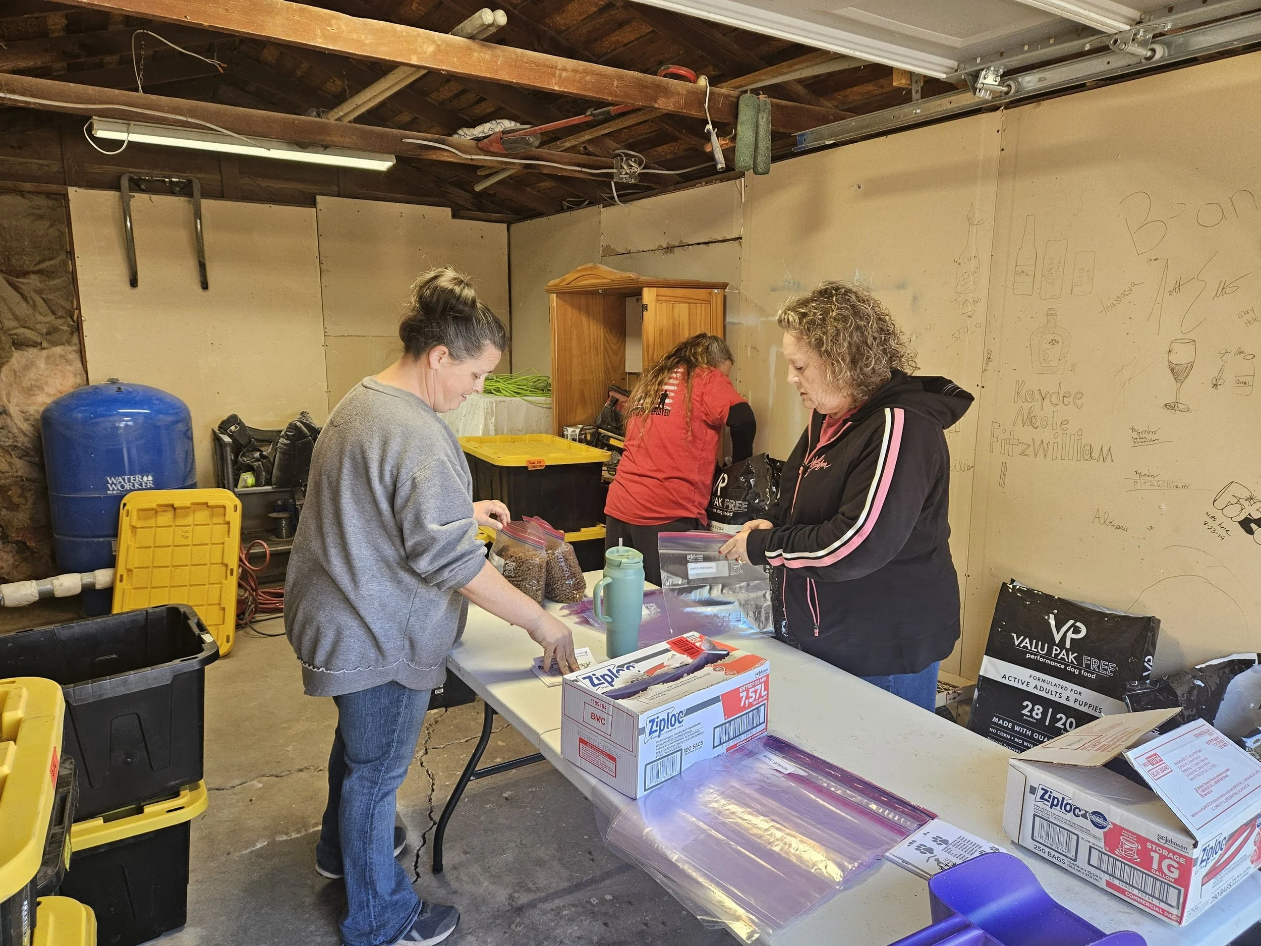 A garage with three people sorting food items into Ziploc bags on tables. The walls have drawings and writing, and there are storage boxes and utility equipment around. The environment suggests a community or volunteer activity.