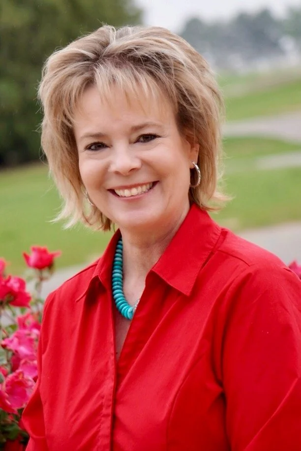 A woman wearing a red shirt and turquoise necklace smiles in a garden setting with pink flowers.