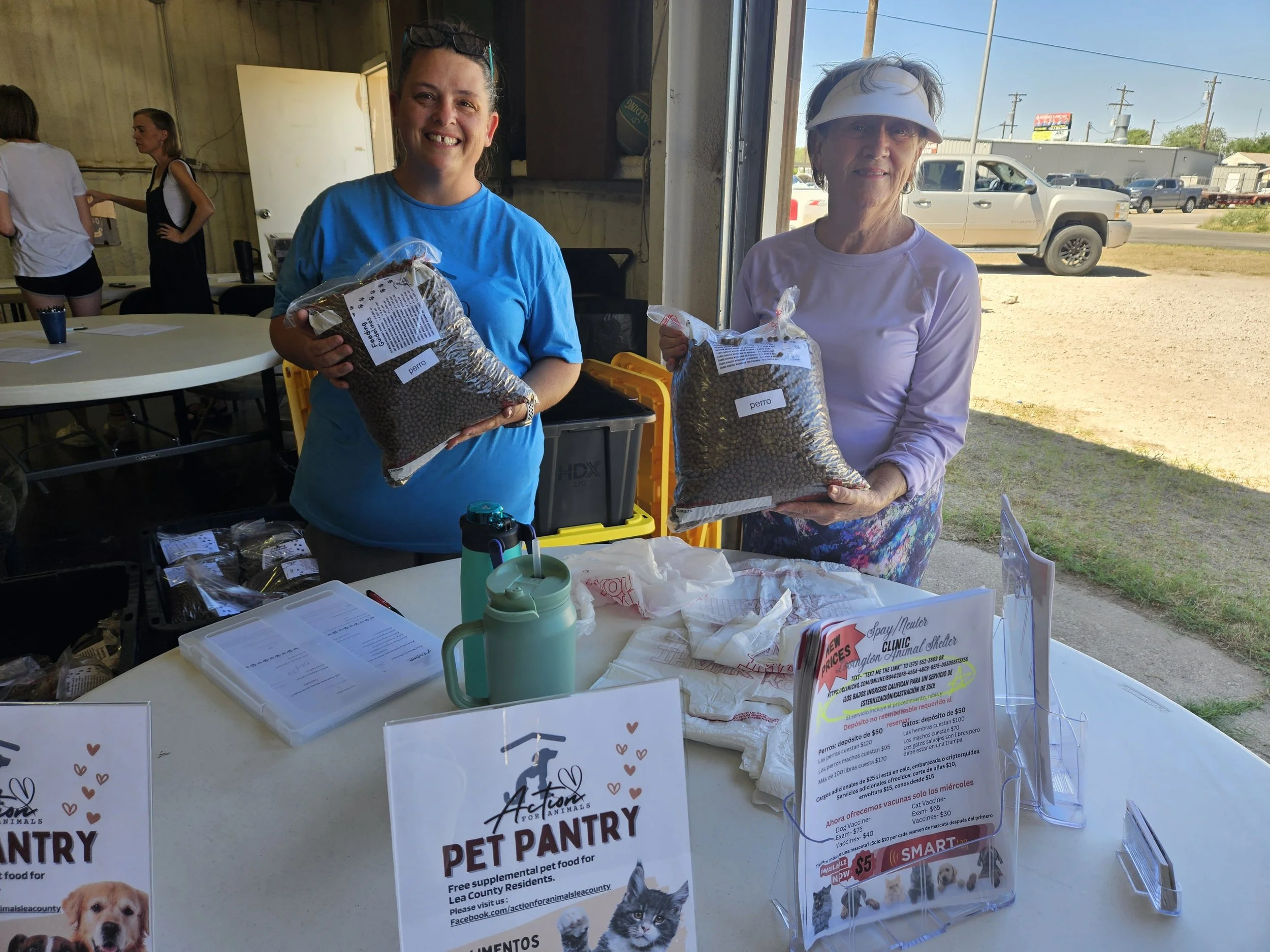 Two women at a pet pantry event holding bags of pet food, with informational signs on a table in front of them.