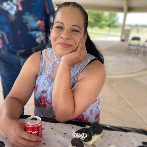 Woman sitting at a table in a floral dress, smiling and resting her chin on her hand, holding a can of Coca-Cola, with a decorated shoe on the table in front of her.