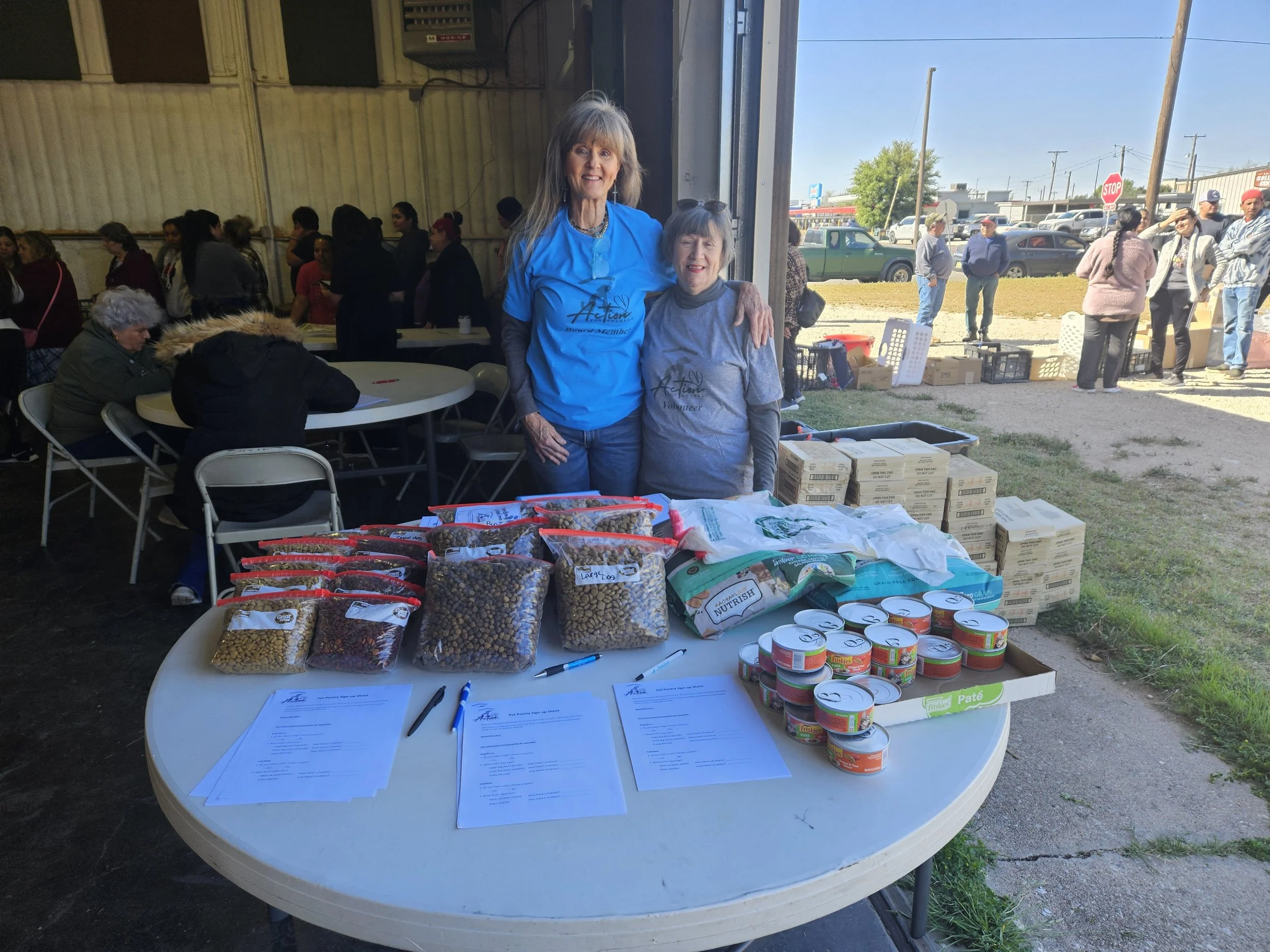 Volunteers at a community food distribution event, featuring tables with bags of dry foods, canned goods, and documents. People are present inside the building and outside, some standing in line. The event appears to be well-attended.