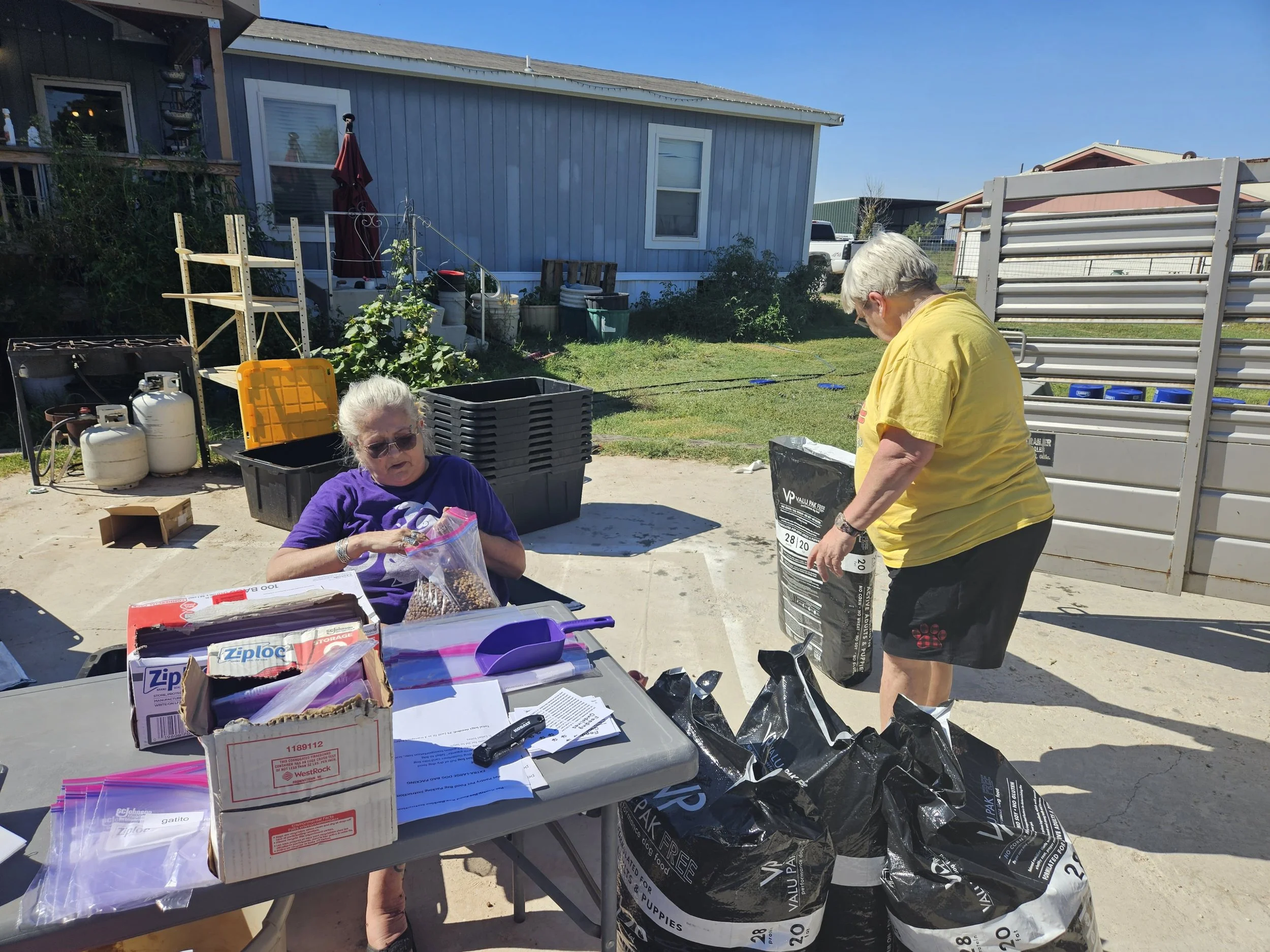 Two women outside near a table. One is sitting, sealing a plastic bag with items; the other is standing, lifting a large bag. There are boxes, bags, and household items around them. A house and yard are in the background.
