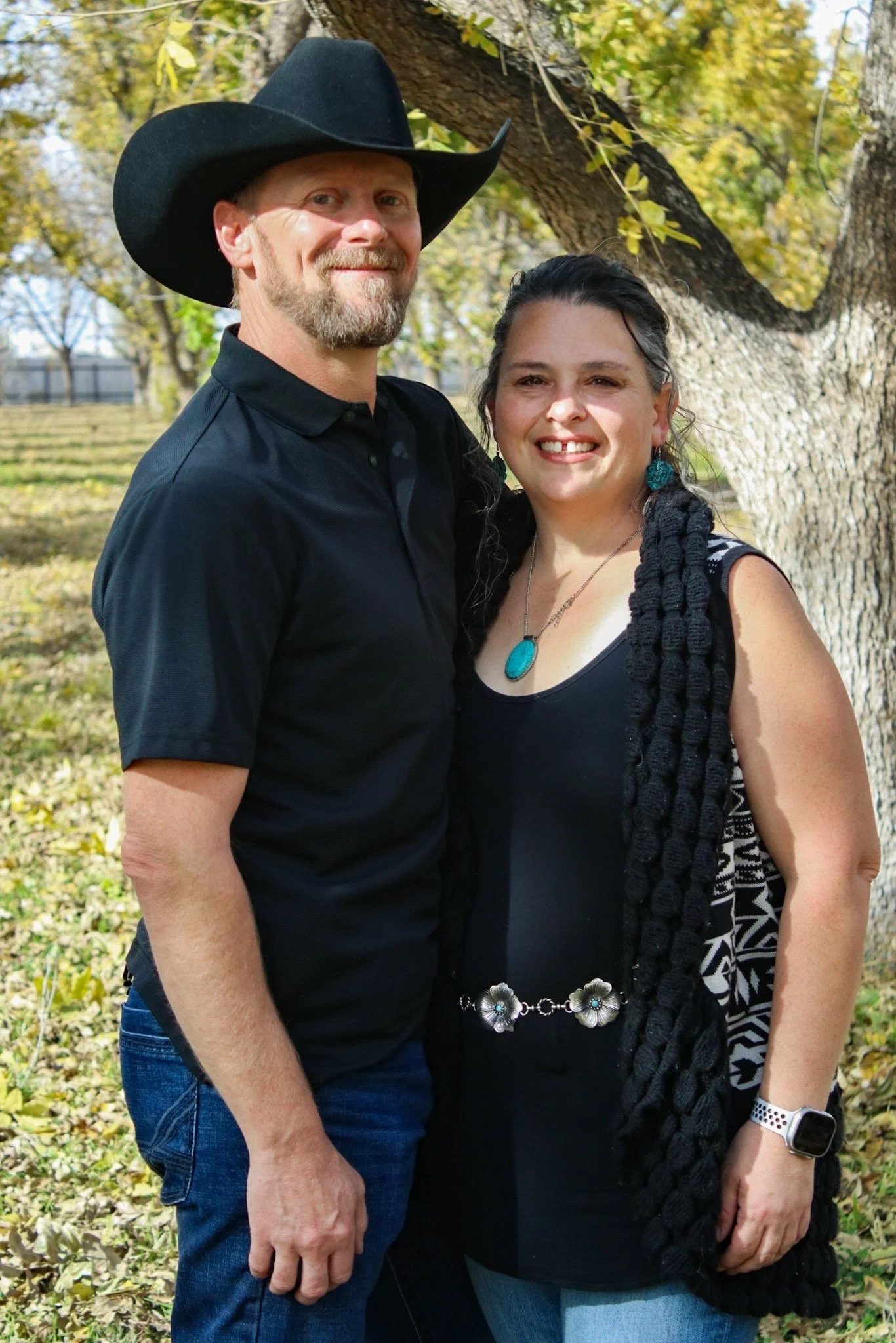A smiling couple, the man wearing a black cowboy hat and shirt, and the woman wearing a black outfit with turquoise jewelry, standing in a leafy park setting.