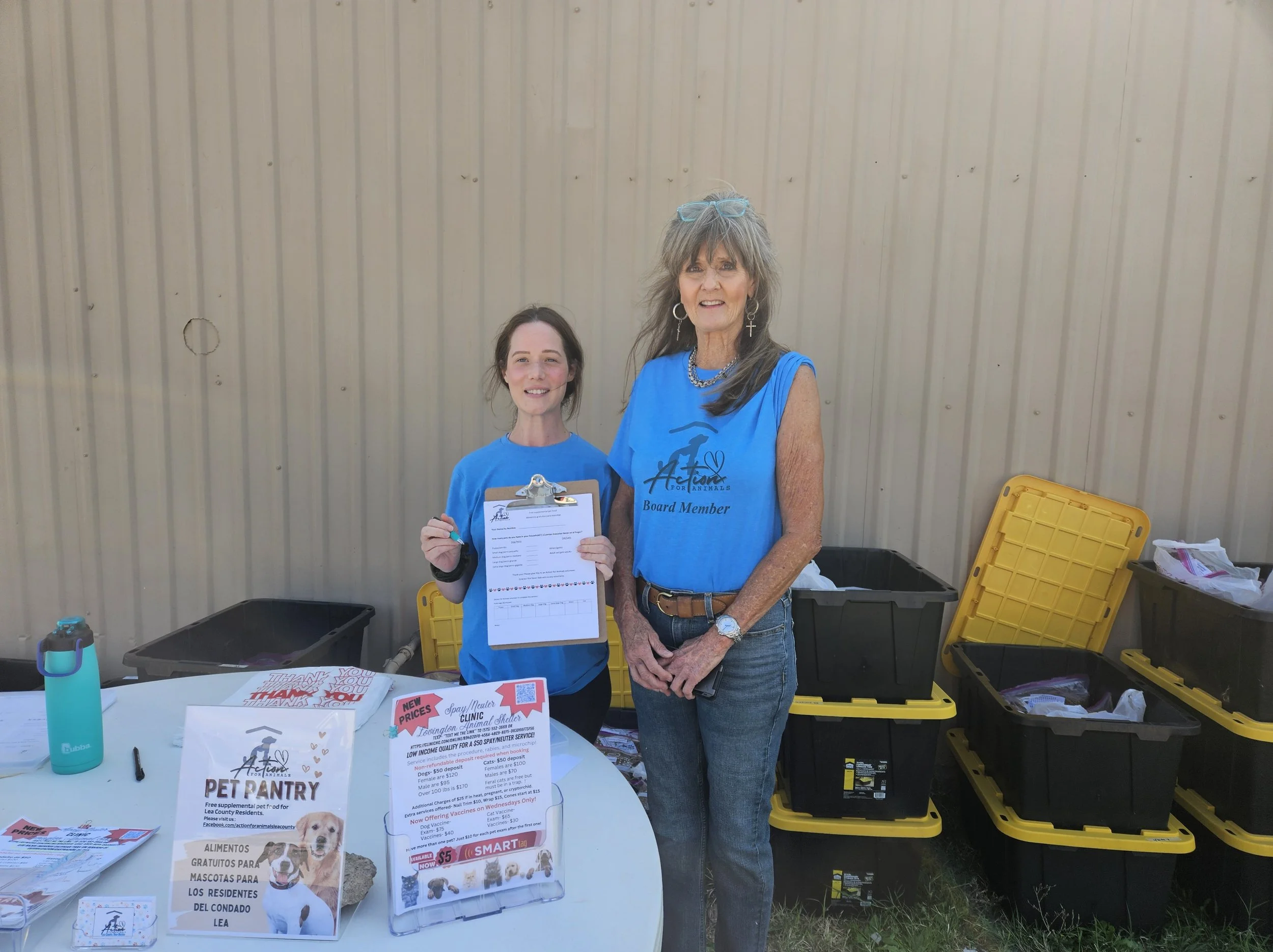 Two women in blue shirts standing outside with a table displaying pet pantry materials, clipboard, and informational flyers.