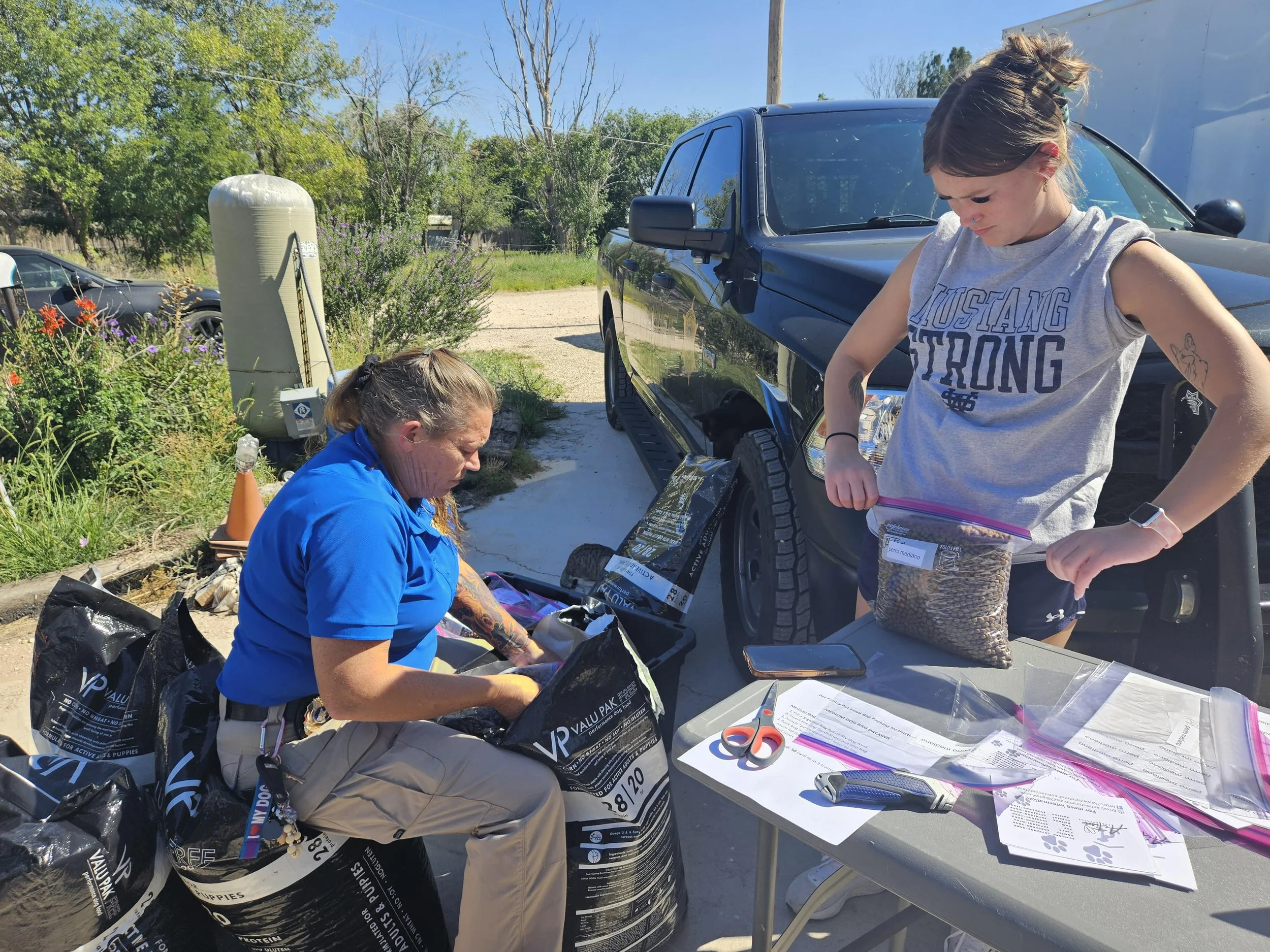 Two people preparing dog food and supplies outdoors next to a black truck. One is wearing a blue shirt and sitting near large black bags labeled 'Valu-Pak,' while the other is wearing a gray 'Mustang Strong' tank top and holding a clear plastic bag o