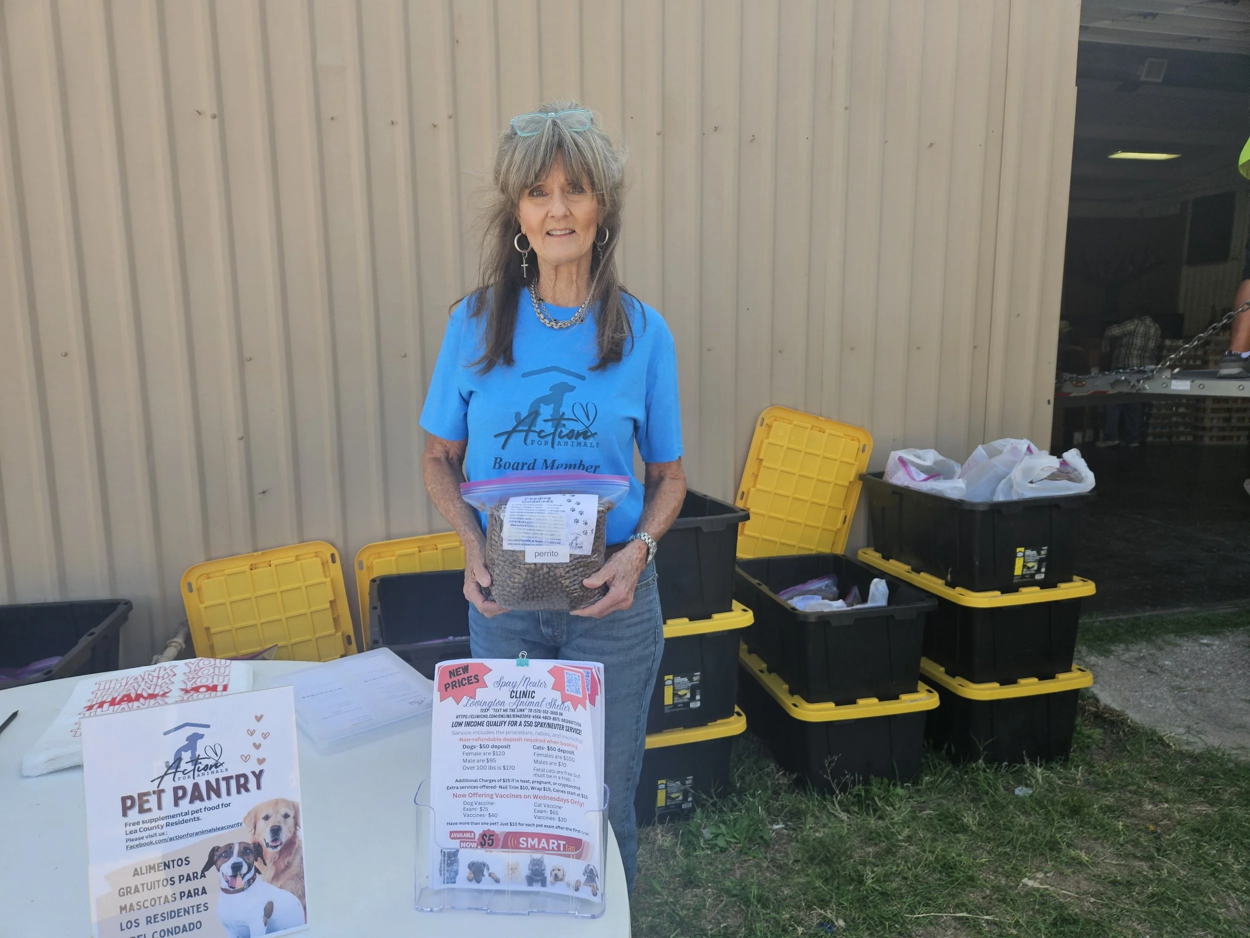 Woman in blue shirt volunteering at an outdoor pet pantry event, holding a bag of pet food. Yellow and black storage bins stacked nearby. Information flyers and a sign about the pet pantry on the table.