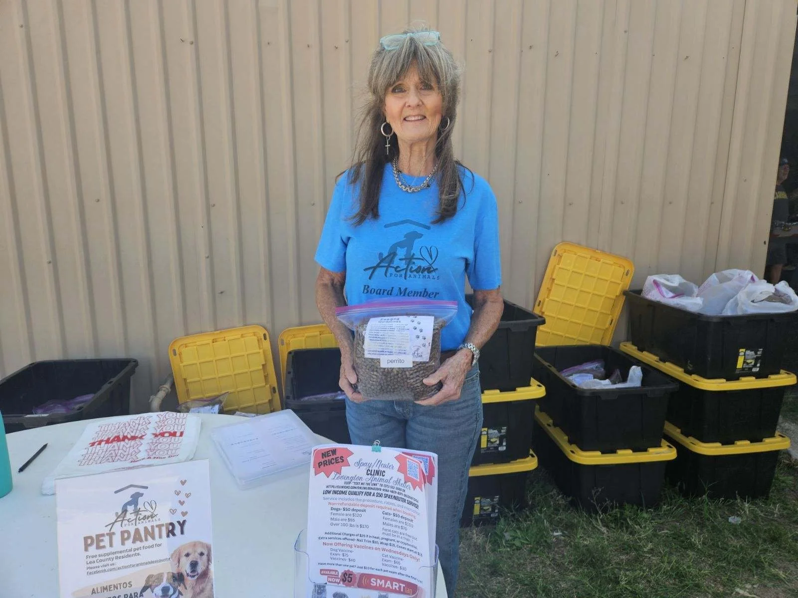 Woman in blue shirt at outdoor pet pantry event, holding a bag of pet food, with informational flyers and storage bins in background.