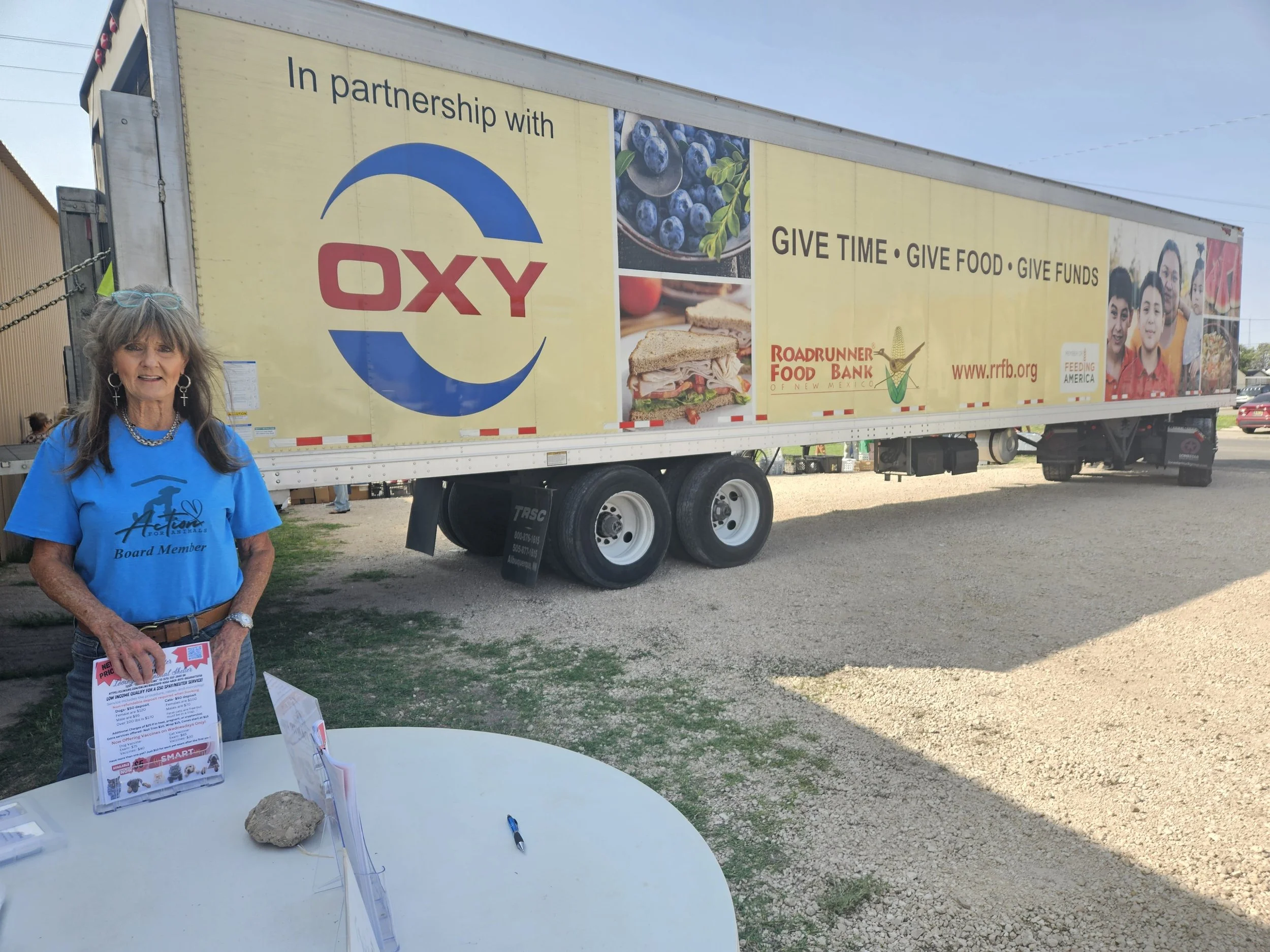 A woman in a blue t-shirt labeled "Board Member" stands in front of a large truck with a banner advertising a partnership. The banner displays "OXY" and "Roadrunner Food Bank of New Mexico" with the message "GIVE TIME • GIVE FOOD • GIVE FUNDS." A tab