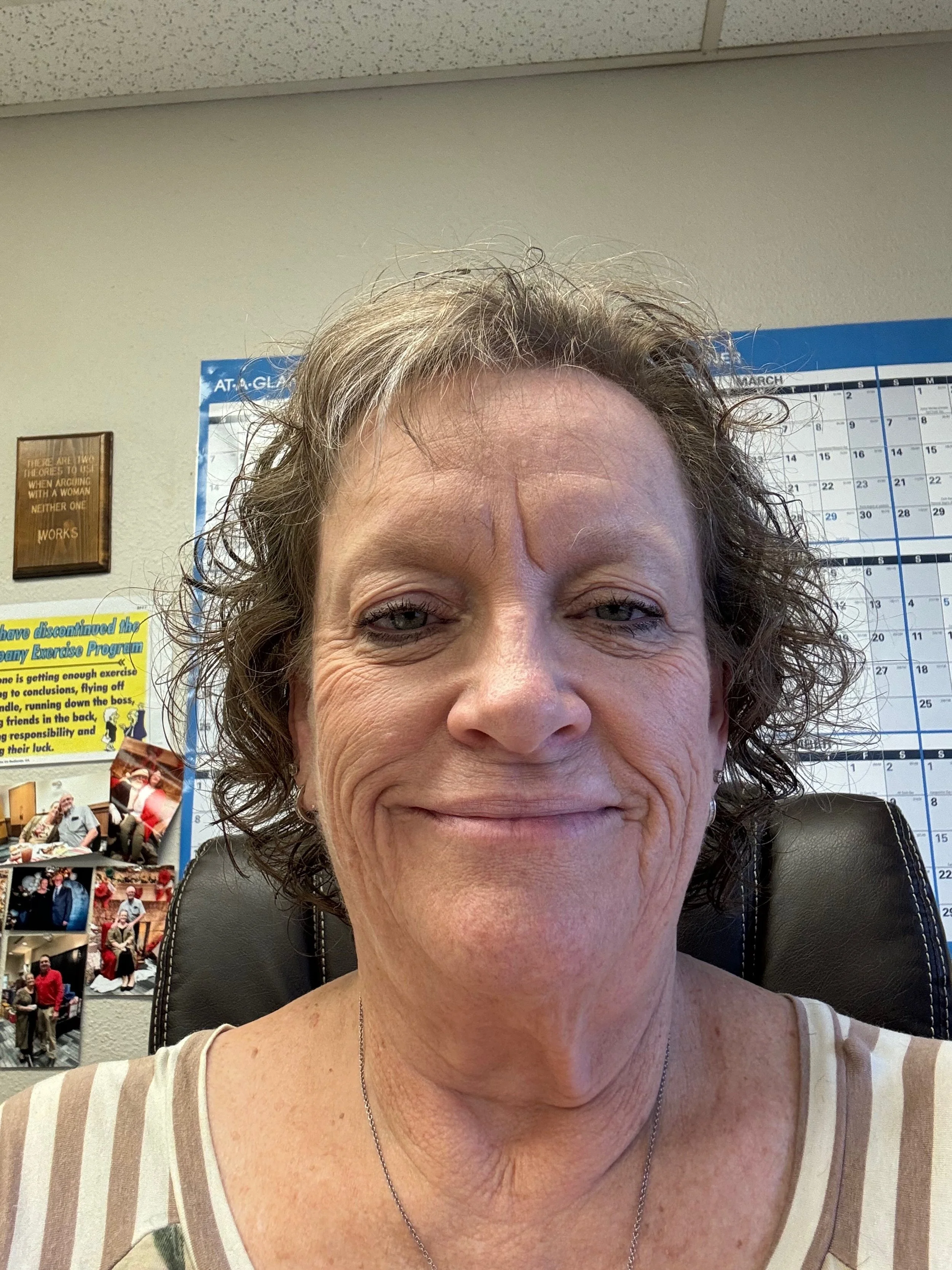 A person with short curly hair is smiling in an office setting. There's a calendar and several posters on the wall in the background.