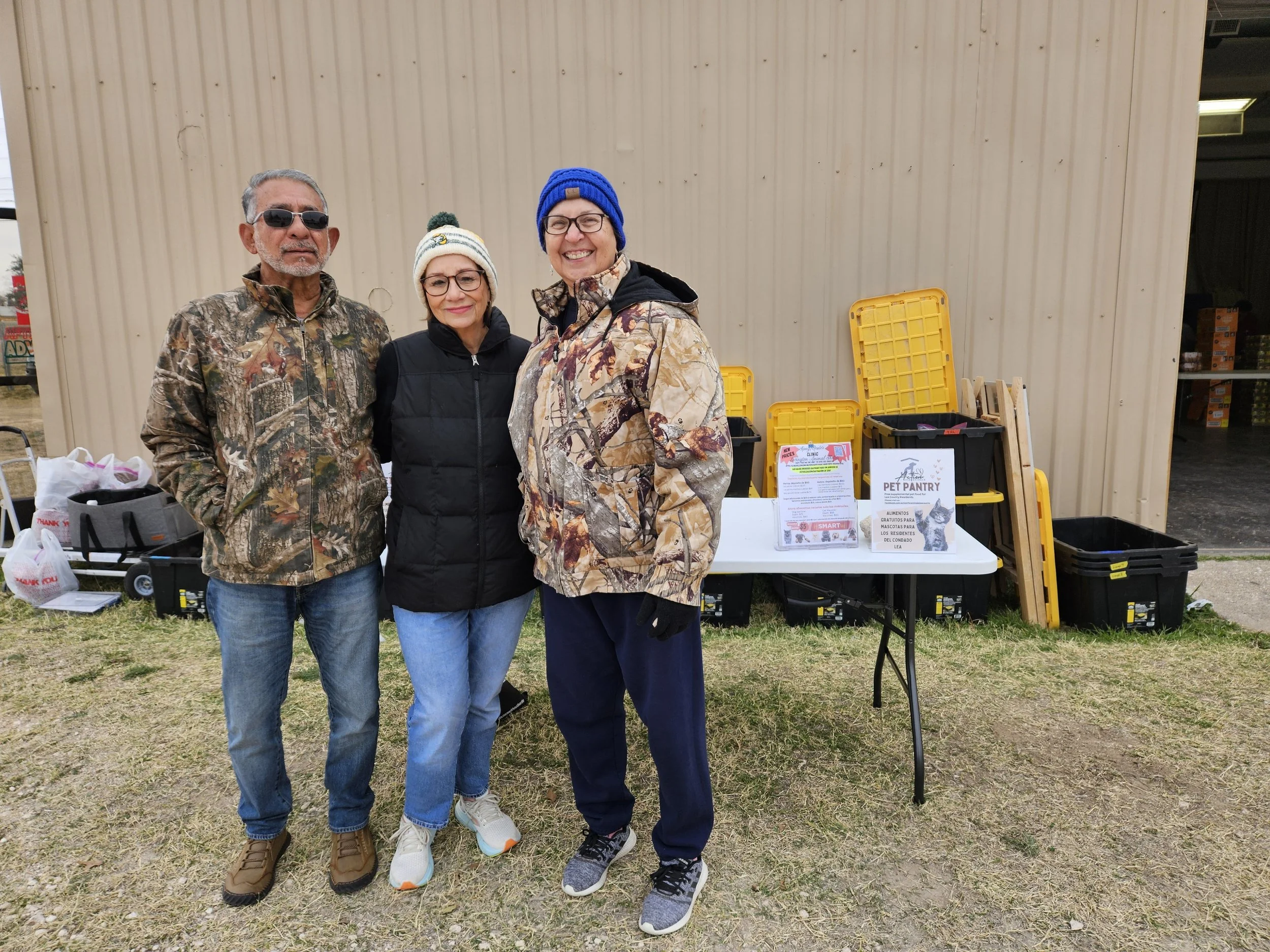 Three people in winter clothing standing outdoors by a table with a pet pantry sign, plastic bins, and supplies.