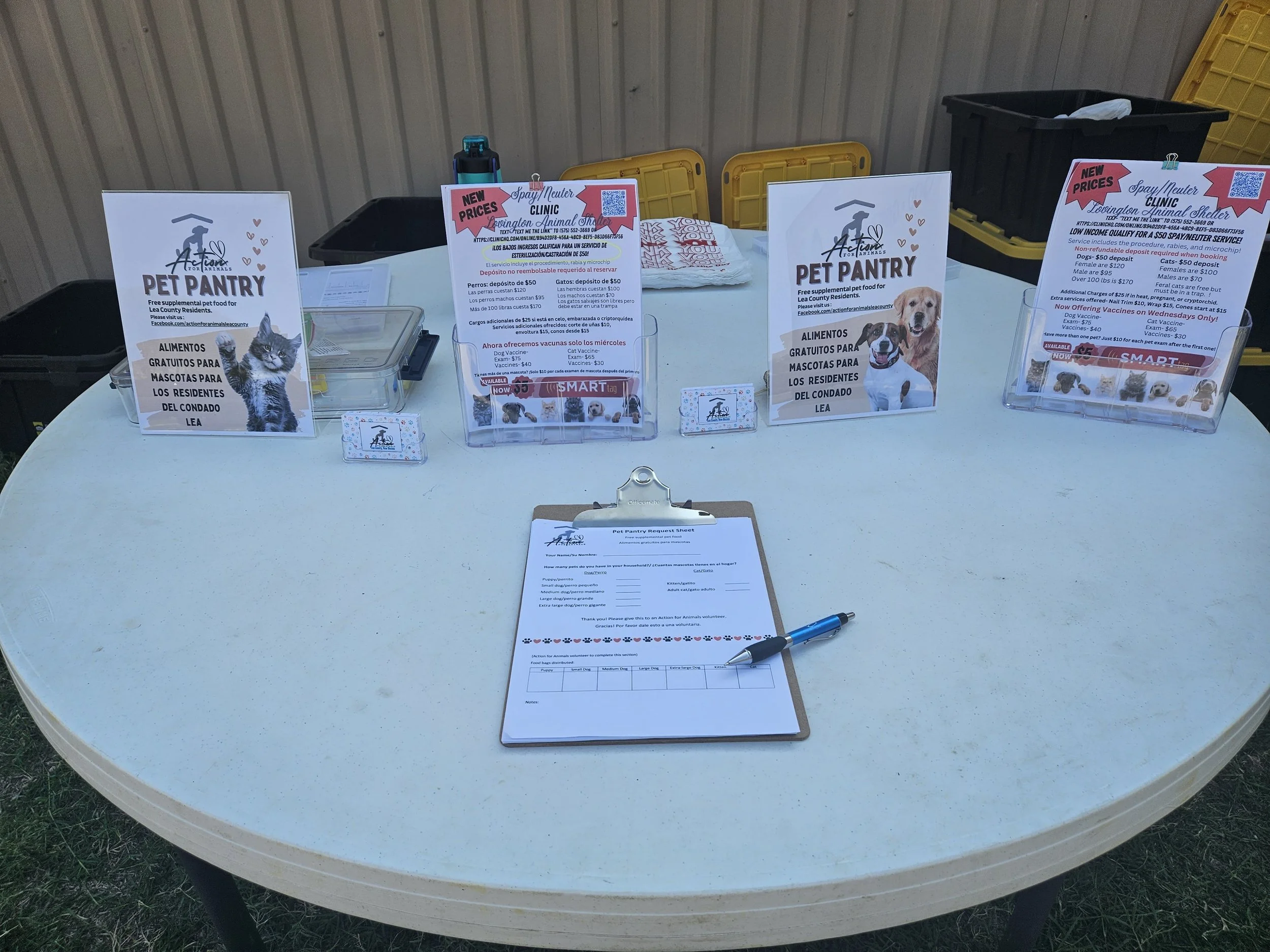 Table display at an animal-related event featuring pet pantry posters in English and Spanish, a clipboard with a form and pen, and informational brochures about pet services.