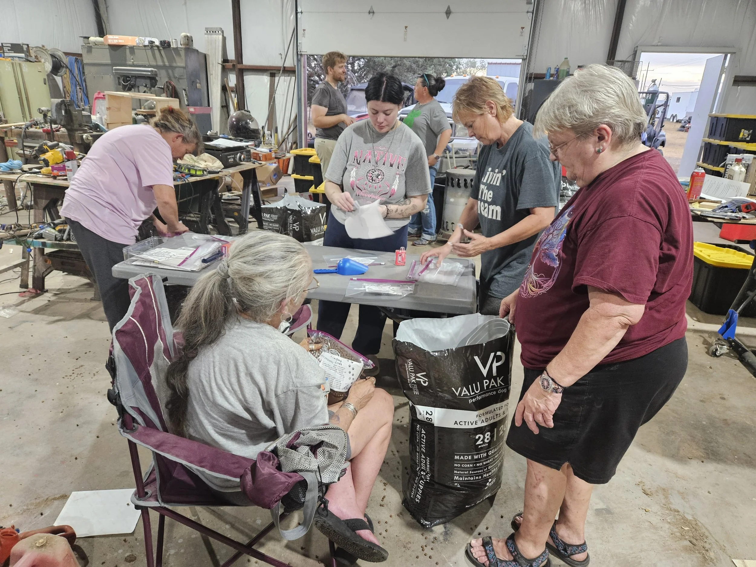 Group of people organizing items in a workshop environment, with a woman in a chair holding papers. Tools and bags scattered around.
